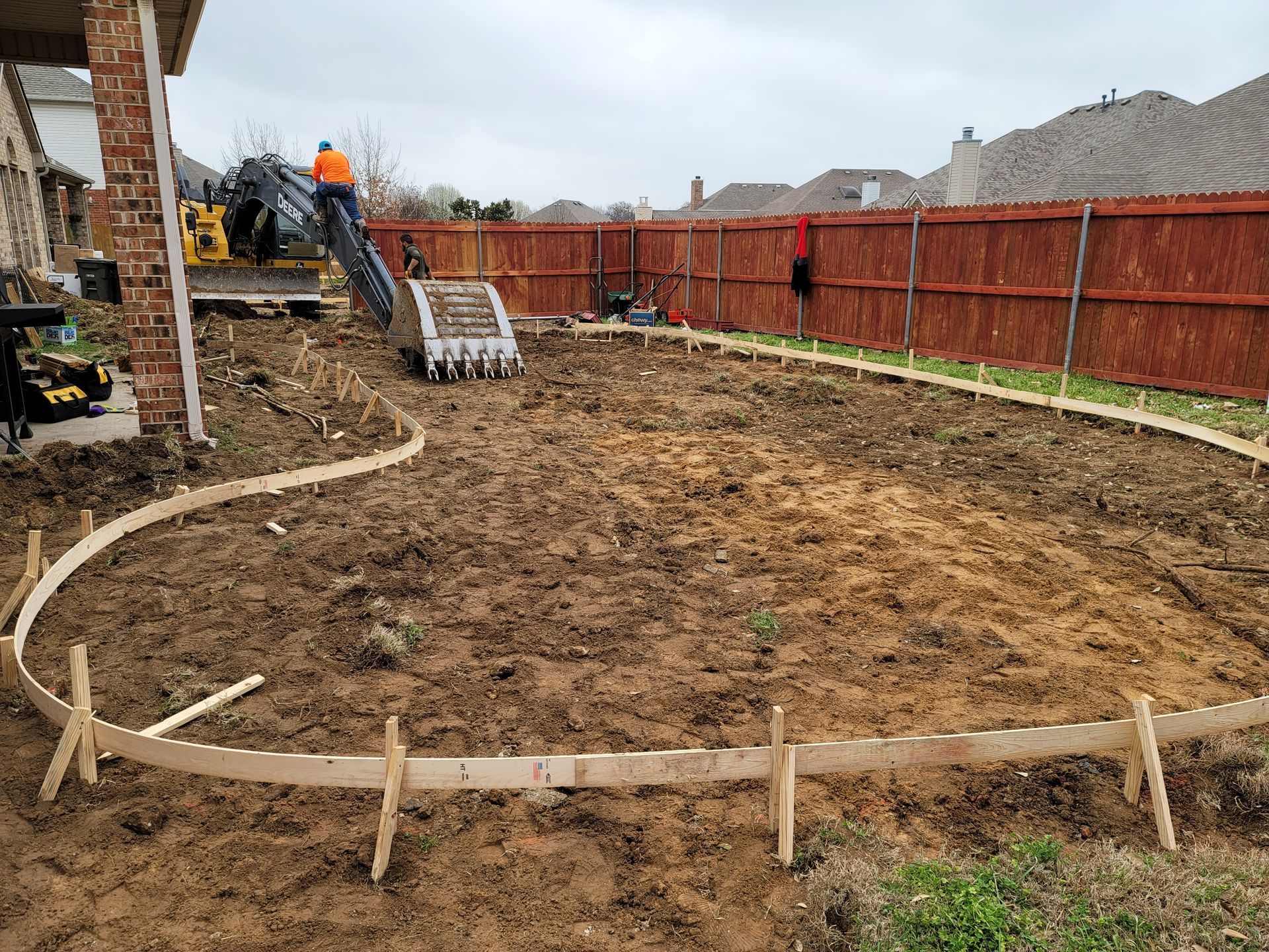 This construction site features flexible wooden forms outlining a large, curved patio or pool deck. A yellow excavator stands in the background near a brick home and a wooden privacy fence.