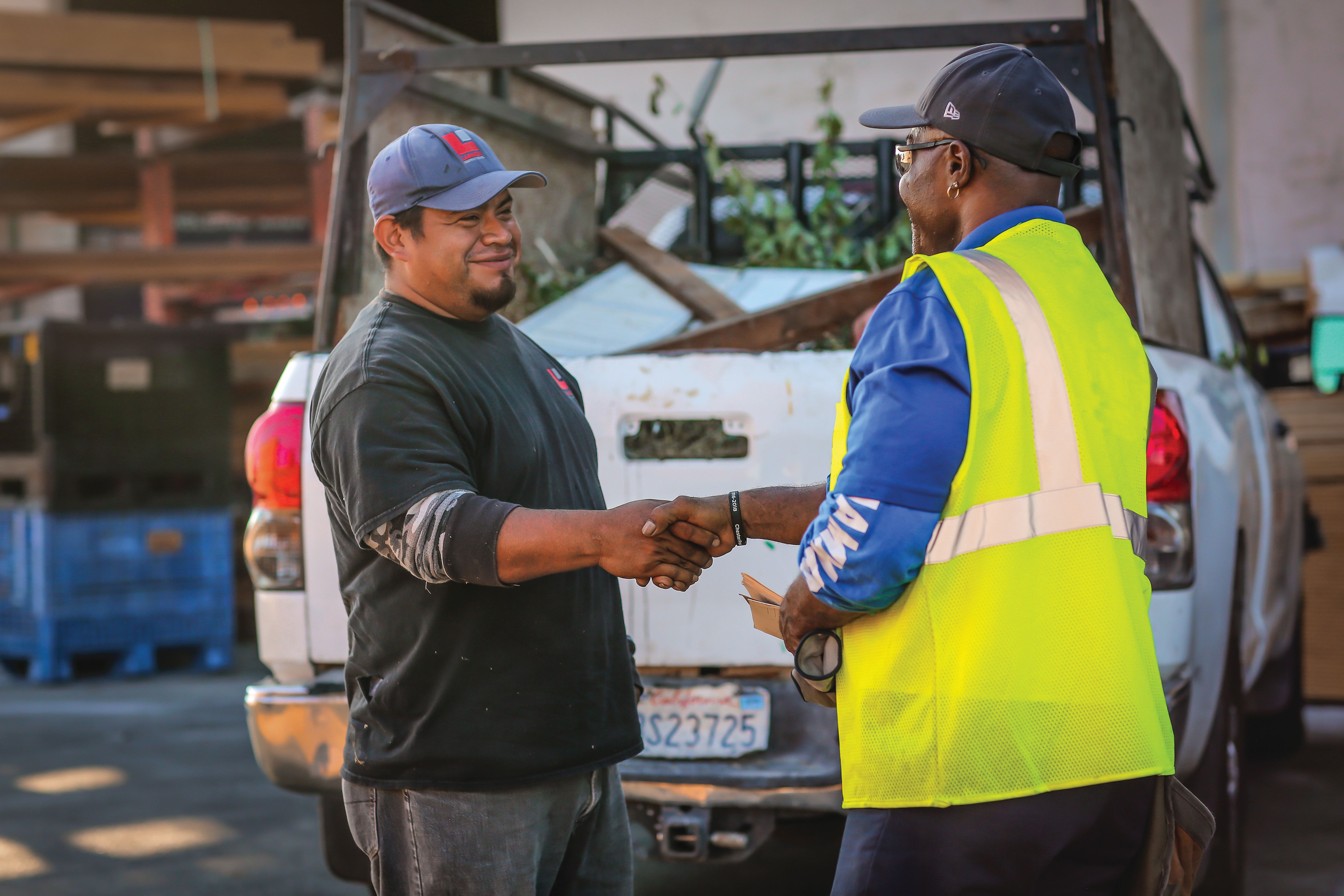 A customer and a Builders FirstSource team member shake hands in a lumber yard next to a pickup truck carrying building materials, representing customer service and jobsite support.