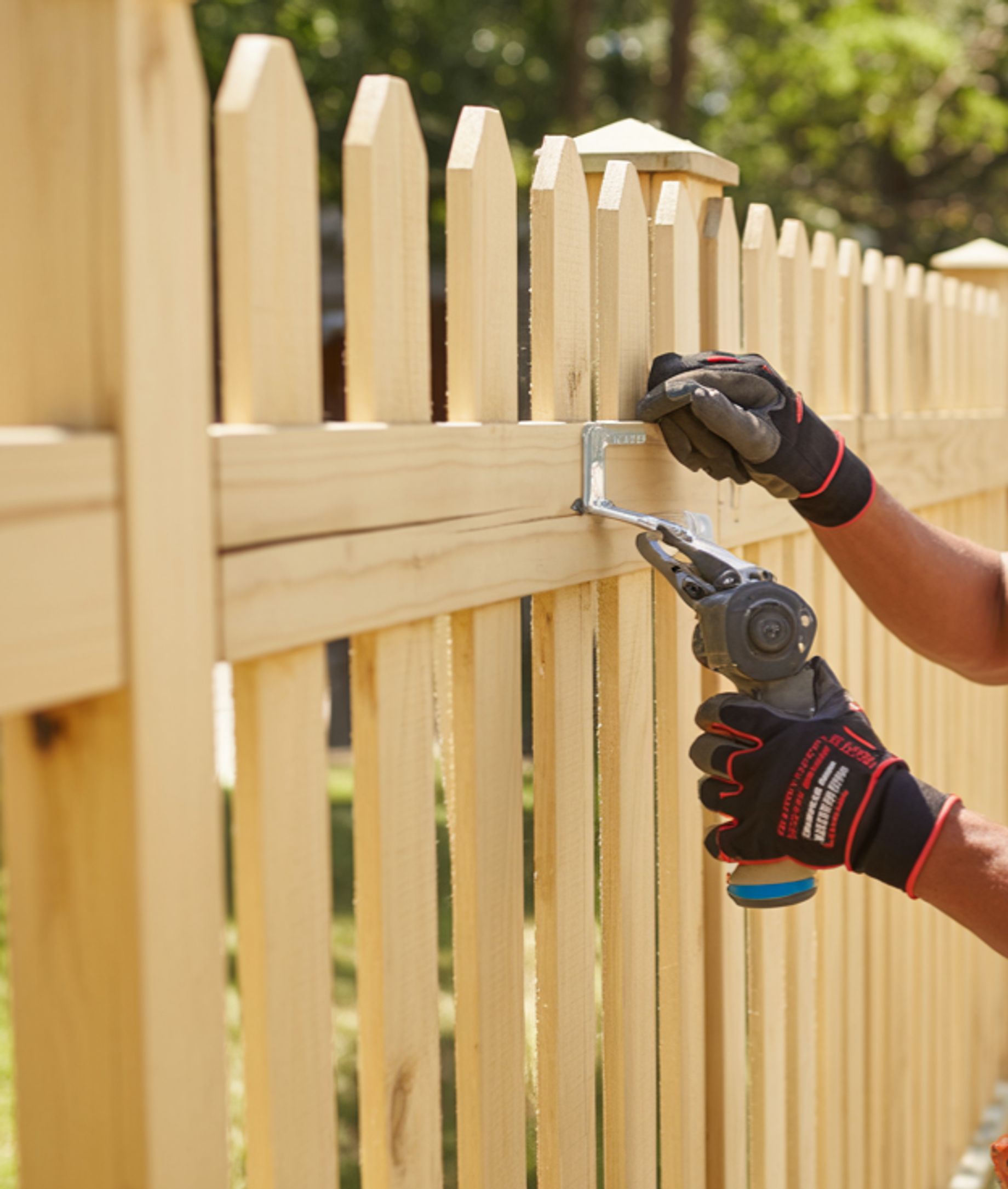 A close-up shot of a person's gloved hands using a tool to attach a bracket to a wooden fence. 