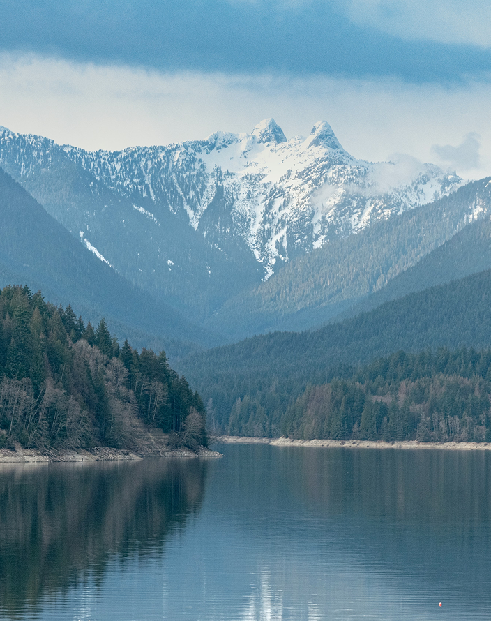 A calm lake surrounded by evergreens beneath a rugged snow-capped mountain.