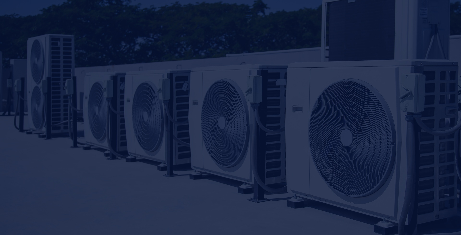 A row of outdoor air conditioning units are lined up on a flat surface with trees in the background.
