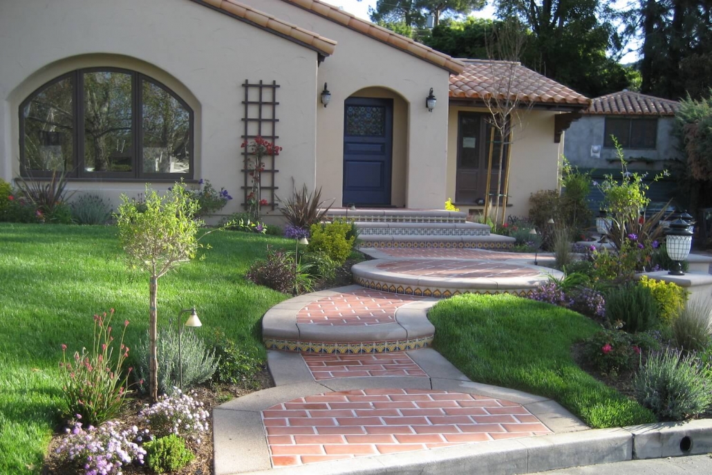 Spanish-style bungalow with terracotta roof, blue door, brick-and-tile pathway, lush lawn, and colorful garden landscaping in warm sunlight.