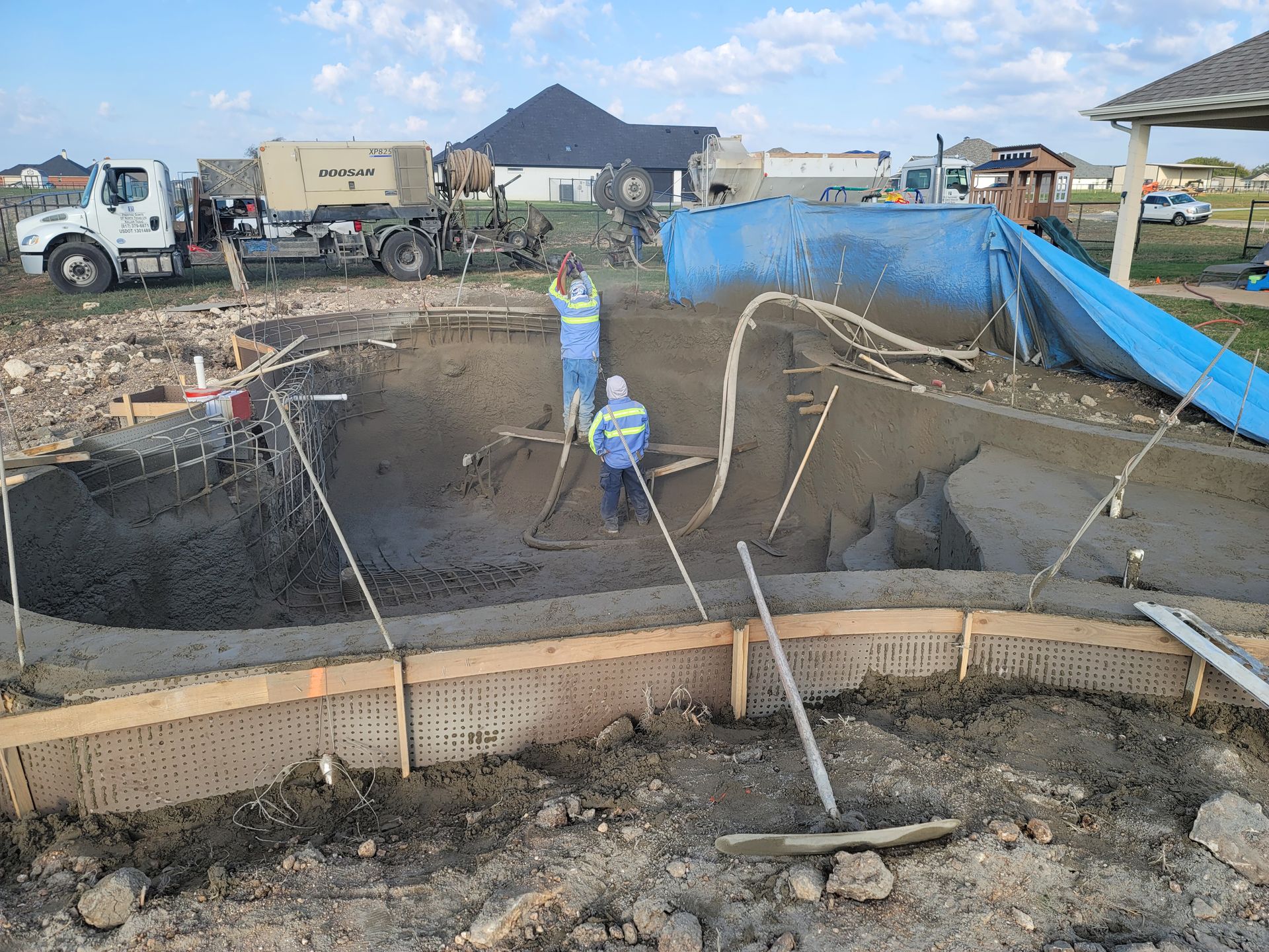 Construction workers in safety gear spray gunite over a steel rebar frame to create the pool shell. Heavy equipment, including a Doosan compressor and several trucks, support the work site.