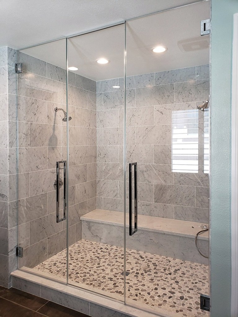 A modern, spacious glass-enclosed shower with marble tile walls, a pebble tile floor, a built-in bench, and chrome fixtures.