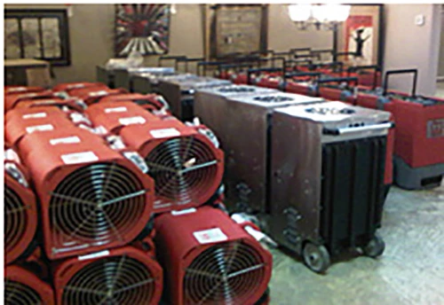 A storage room filled with professional-grade restoration equipment, including stacks of red air movers and industrial dehumidifiers on wheels. The equipment is neatly organized, ready for deployment in water damage restoration projects.