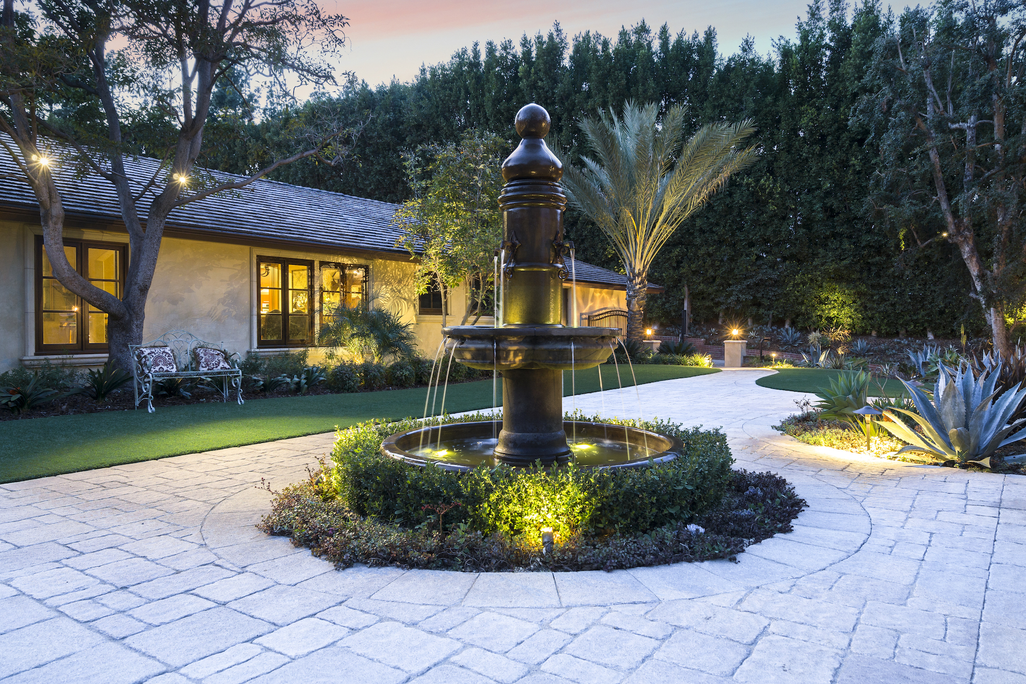Elegant courtyard at dusk with a tiered black fountain, glowing garden lights, stone paving, palms, agave, and a warmly lit home.