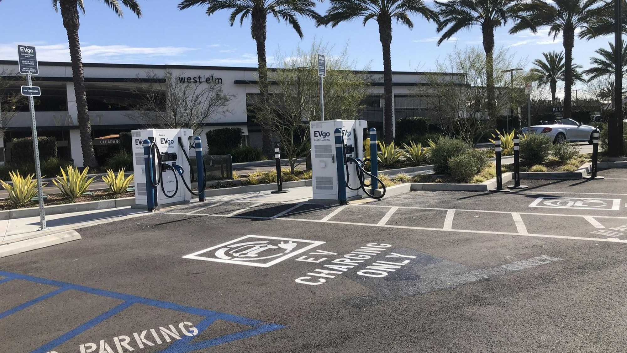 Several EVgo fast charging units installed in marked parking spaces at a public charging station, with EVgo logos on the chargers, protective bollards around each unit.