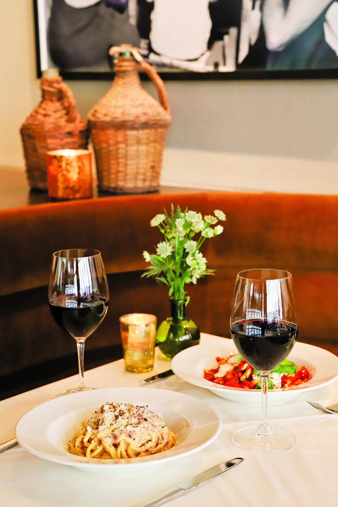 A dinner scene with two pasta dishes and red wine on a white linen table, set against a plush orange banquette and vintage-style decor.