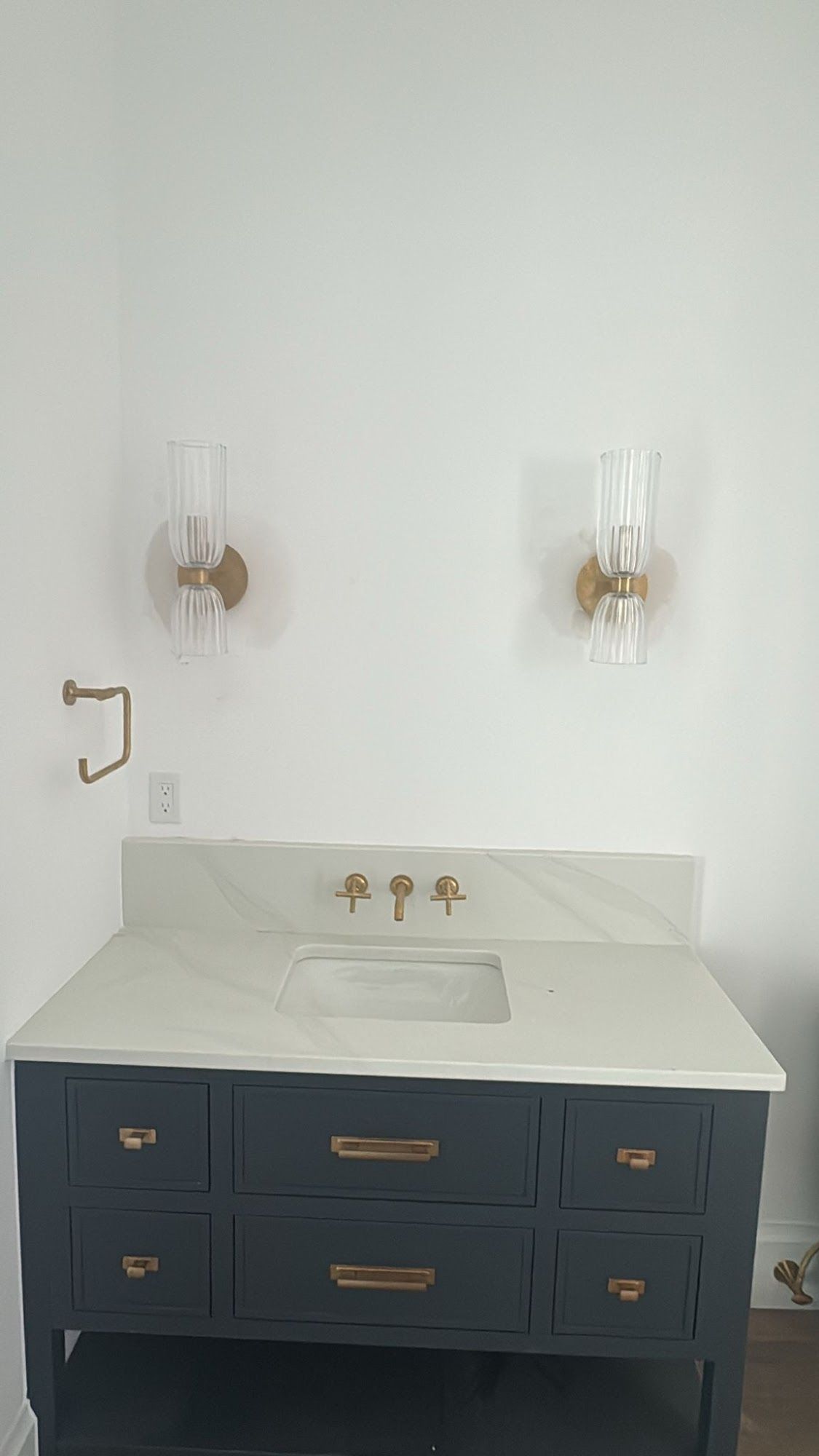 Modern bathroom vanity featuring a dark blue cabinet with brass handles, white countertop sink, gold wall-mounted faucet, and two decorative wall sconces above, creating a clean, minimalist interior design aesthetic.