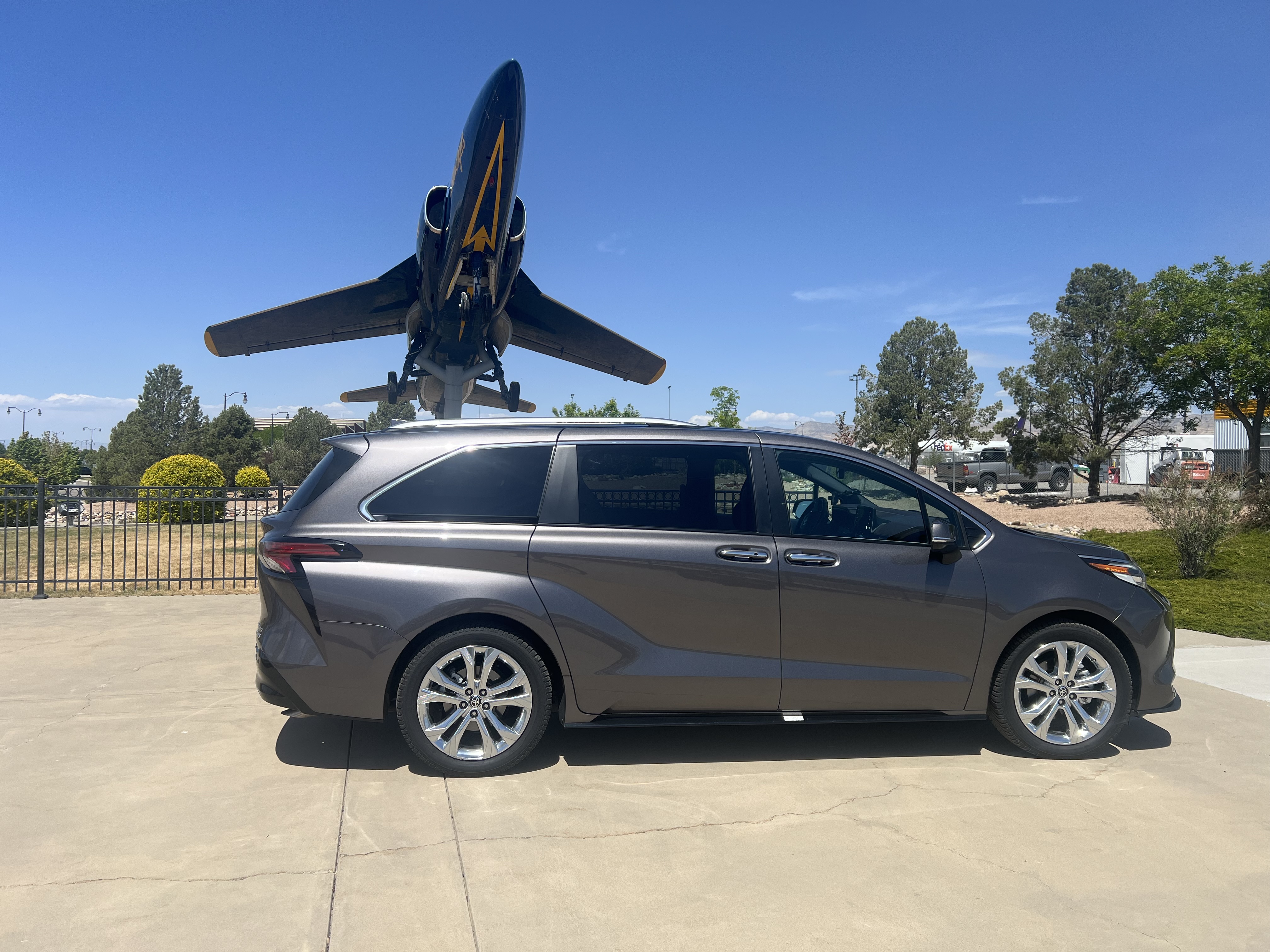 A dark gray minivan is parked in front of a display of an airplane. The airplane is dark blue with yellow accents and is mounted on a pole, angled upwards. The minivan is positioned to the right of the airplane, and the background shows trees, a fence, and some buildings in the distance under a clear blue sky.