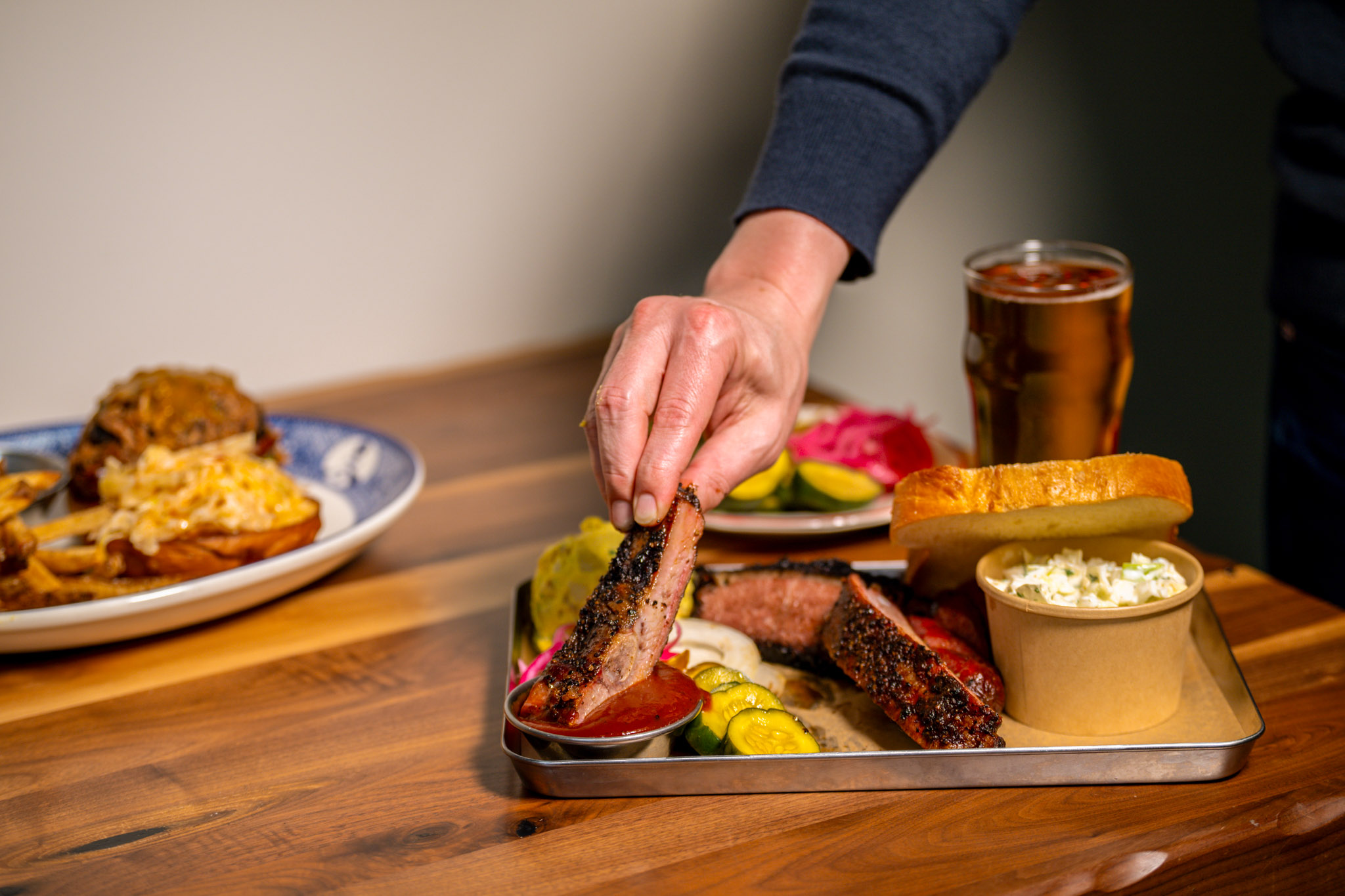 A hand reaches in to pick up a glazed spare rib from a metal BBQ tray loaded with sliced smoked brisket, bread and butter pickles, pickled red onions, white bread, coleslaw, and Joe's BBQ sauce, with a second plate and cold beer visible in the background.