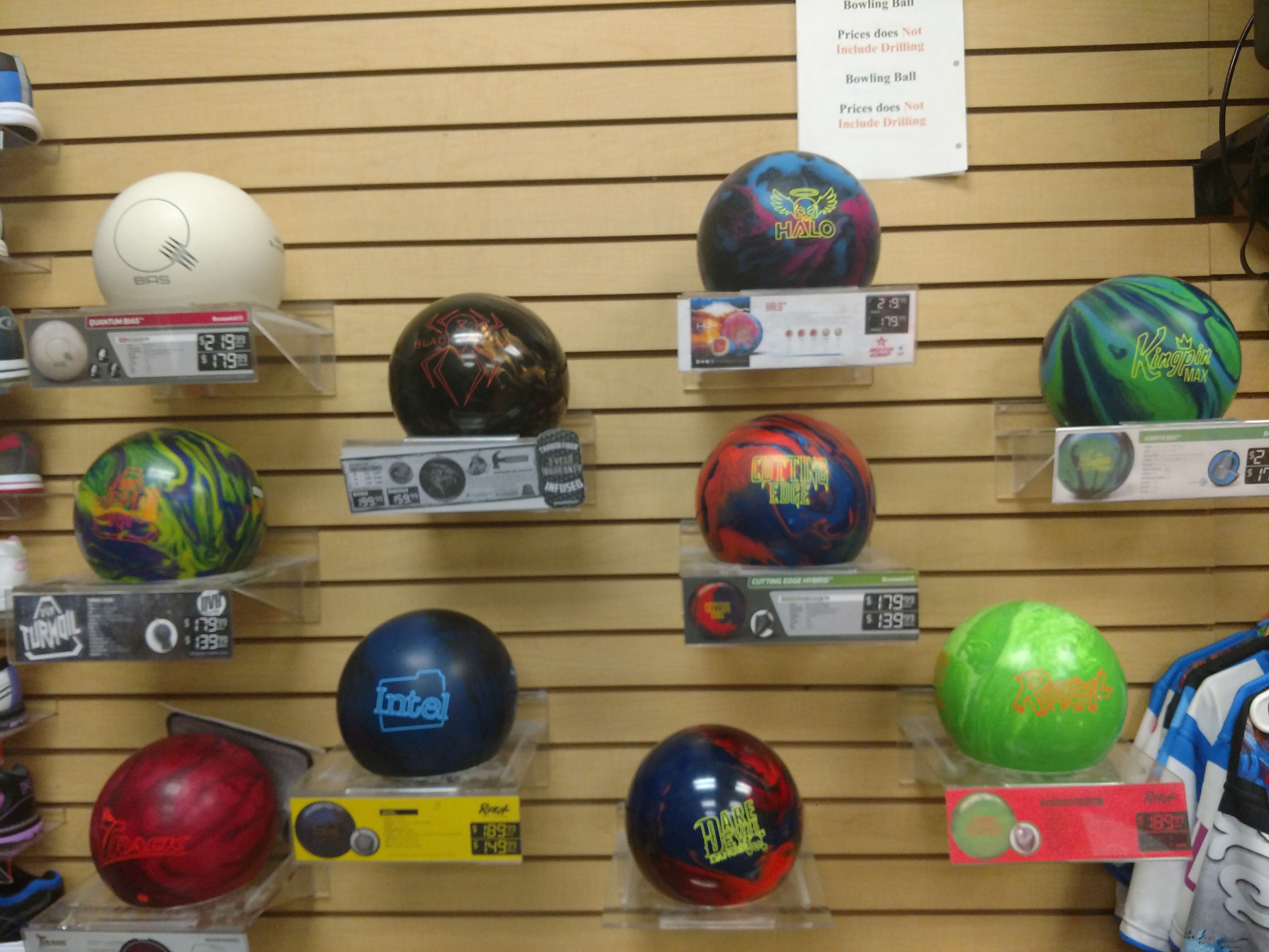 A wall display shows an assortment of bowling balls in various colors and designs. Each ball is on a shelf, and some have price tags or product information displayed below them. The background is a light-colored wood-paneled wall with horizontal slats. Several shirts are also visible on the right side of the image, suggesting this is a retail environment for sporting goods.