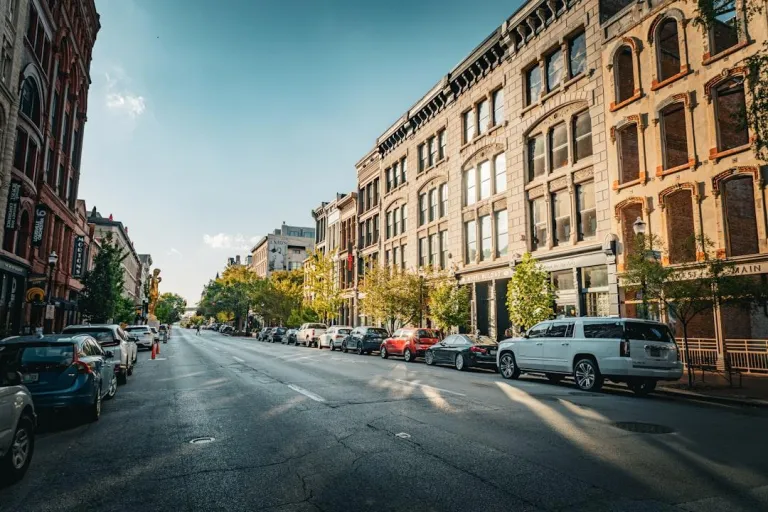 A sun-drenched street lined with historic brick buildings and parked cars basks in golden hour light. The road, asphalted and worn, stretches into the distance, leading to a visible statue and trees. Cars of various colors and models are parked along both sides of the street, with a white SUV prominently featured on the right.
