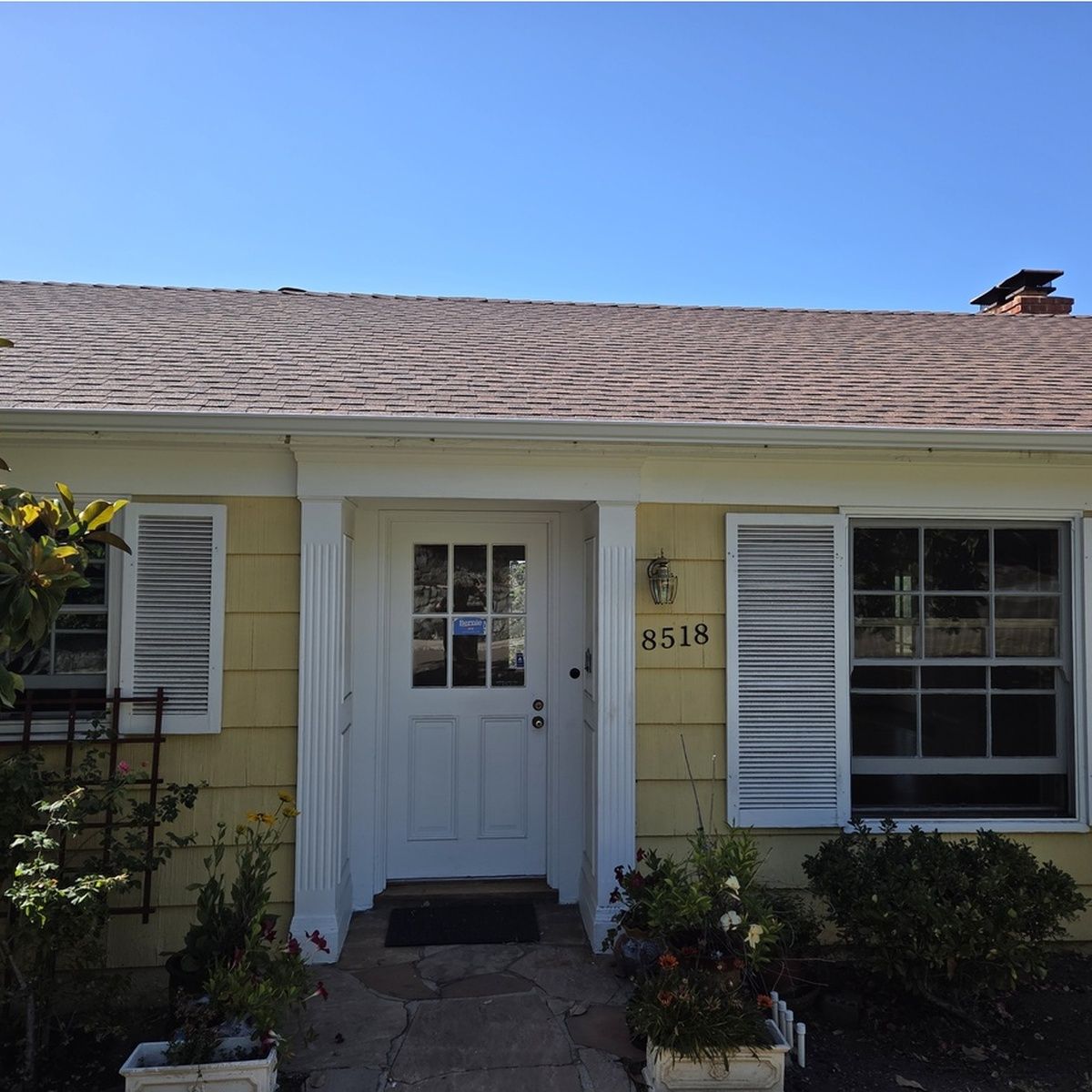 Front exterior of a charming yellow house featuring a freshly shingled roof, white entry door with paneled windows, white shutters, and potted plants framing the stone walkway.