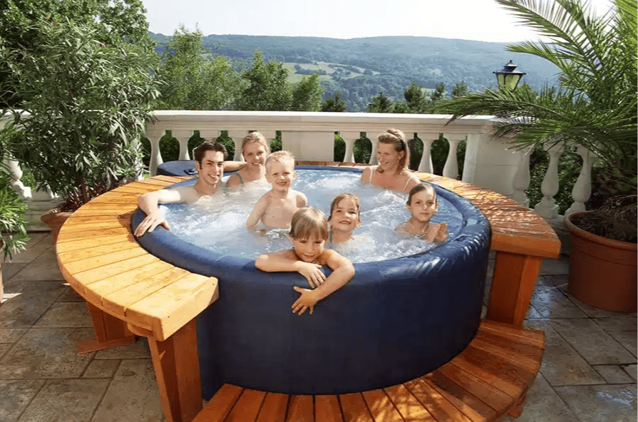 A Softub Resort 300 portable hot tub in navy blue is shown fully activated with jets running on a stone tile terrace with a panoramic view of rolling green hills. The tub is surrounded by a custom curved cedar or redwood deck surround, enhancing its aesthetic appeal and providing a place to sit. A family of seven &mdash; two adults and five children of varying ages &mdash; are all enjoying the spa together, conveying its large capacity and family-friendly design. The terrace setting features white balustrade railings, tropical potted palms, a lantern post, and lush greenery, giving the scene a resort-like feel on a bright summer day.