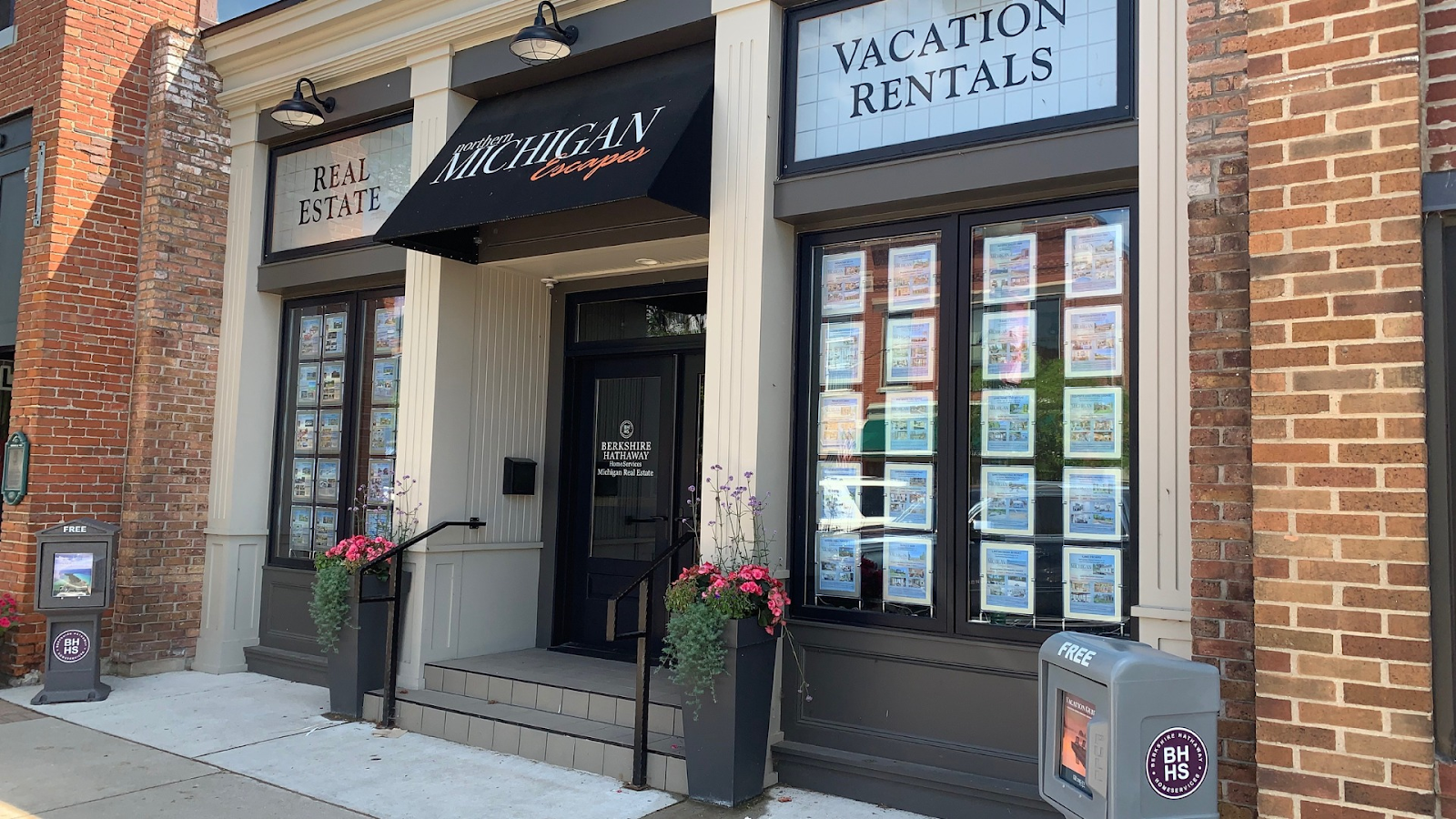 A real estate office with "Northern Michigan Escapes" and "Vacation Rentals" signage is pictured. The office windows display numerous real estate listings. A "FREE" sign is visible on the left, and a brochure dispenser with the "BH HS" logo is on the right.