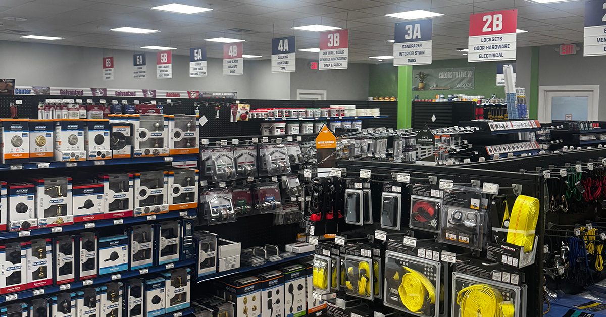 Wide horizontal view of two Builders FirstSource aisles. The foreground shelf displays rows of Rust-Oleum spray paint and primer in multiple colors, while the adjacent aisle contains packaged saw blades, drill bits, fasteners and tools. Overhead signs label sections 4B through 6B for saw blades, drill bits, staples, fasteners, boxed nails, flashing, framing connectors and power tools.