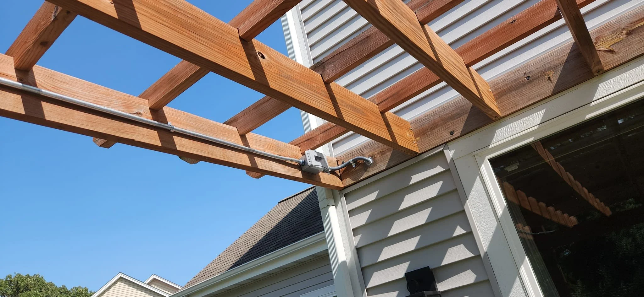 A wooden pergola is attached to the side of a house, with electrical conduit running along one of the beams and connected to a weatherproof outlet box. The house has light gray siding and a white trim around a window. The sky is clear blue.