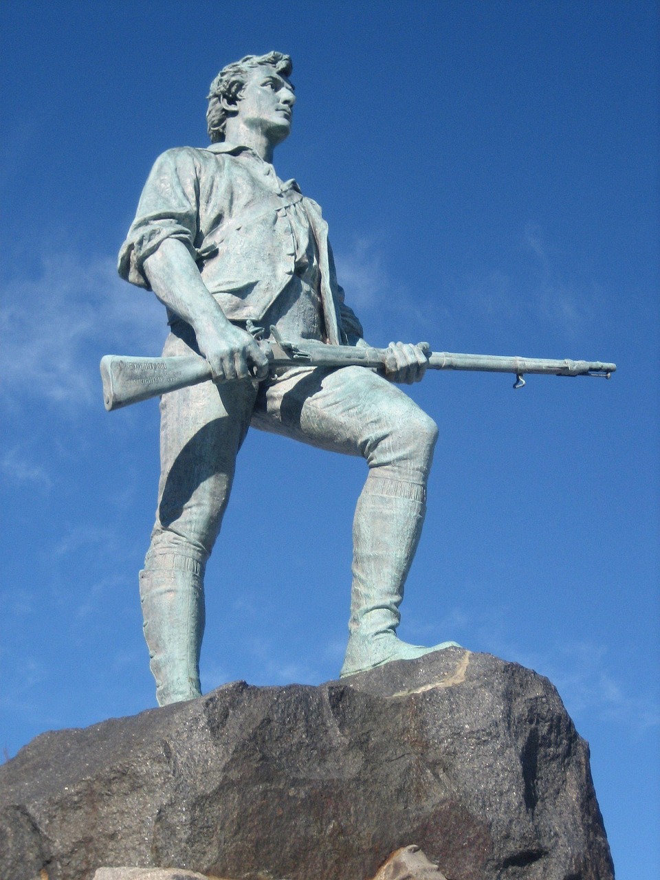 A bronze statue of a Minute Man stands atop a rocky outcrop under a bright blue sky. The statue depicts a determined man in colonial-era attire, holding a musket. He wears a loose-fitting shirt, a vest, breeches, and tall boots. His gaze is directed to the right, suggesting alertness and readiness. The statue appears weathered, with a greenish patina indicative of aged bronze.