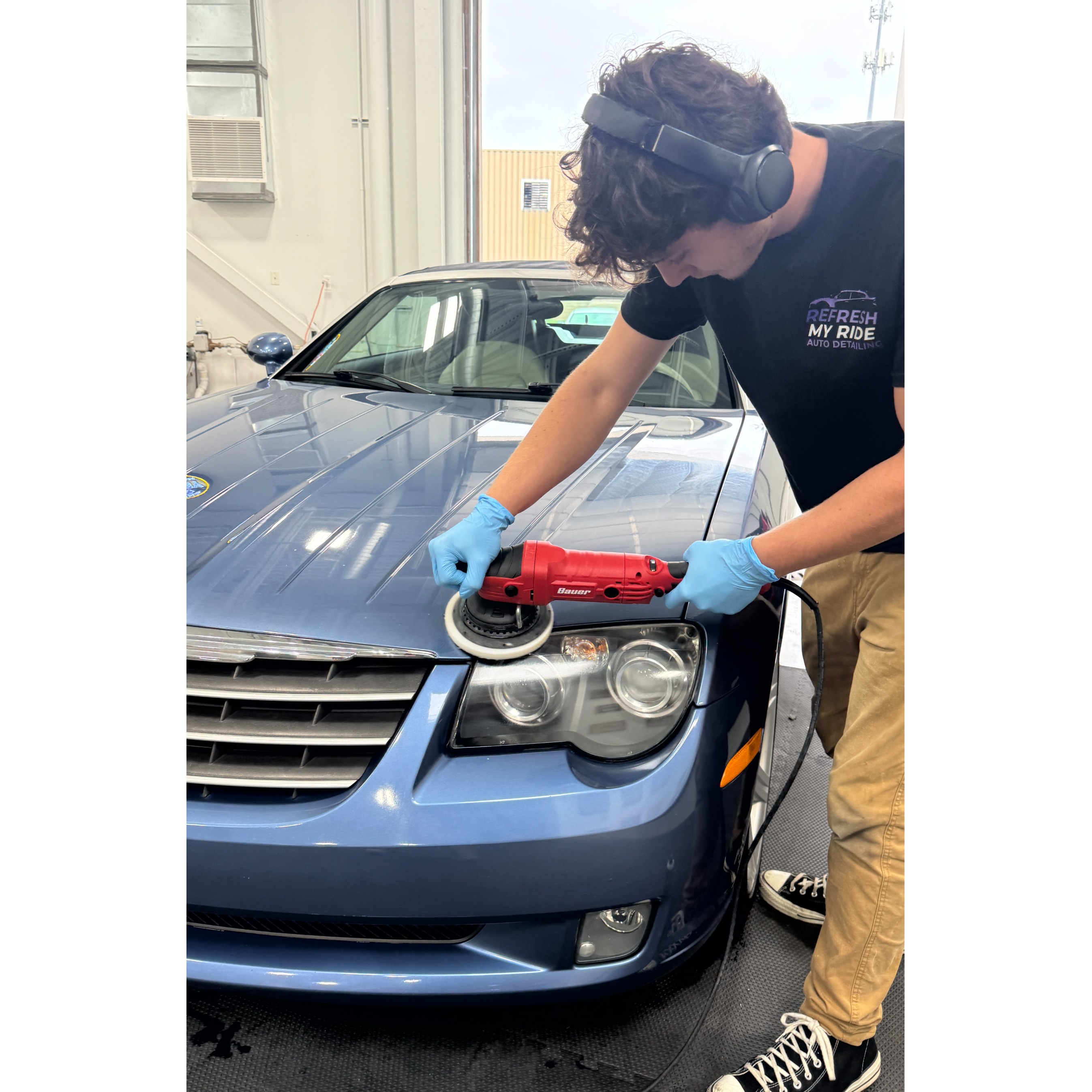 A professional technician at Refresh My Ride performs high-end paint correction on a blue sedan at their specialized auto detailing shop. Wearing protective gloves and a company uniform, the detailer uses a specialized machine polisher to eliminate swirl marks and surface defects, restoring the vehicle's paint to a brilliant, deep shine. This level of precision is an essential step in the ceramic coating process, ensuring clients in Macomb and Oakland Counties receive the highest standard of vehicle protection and aesthetic restoration.