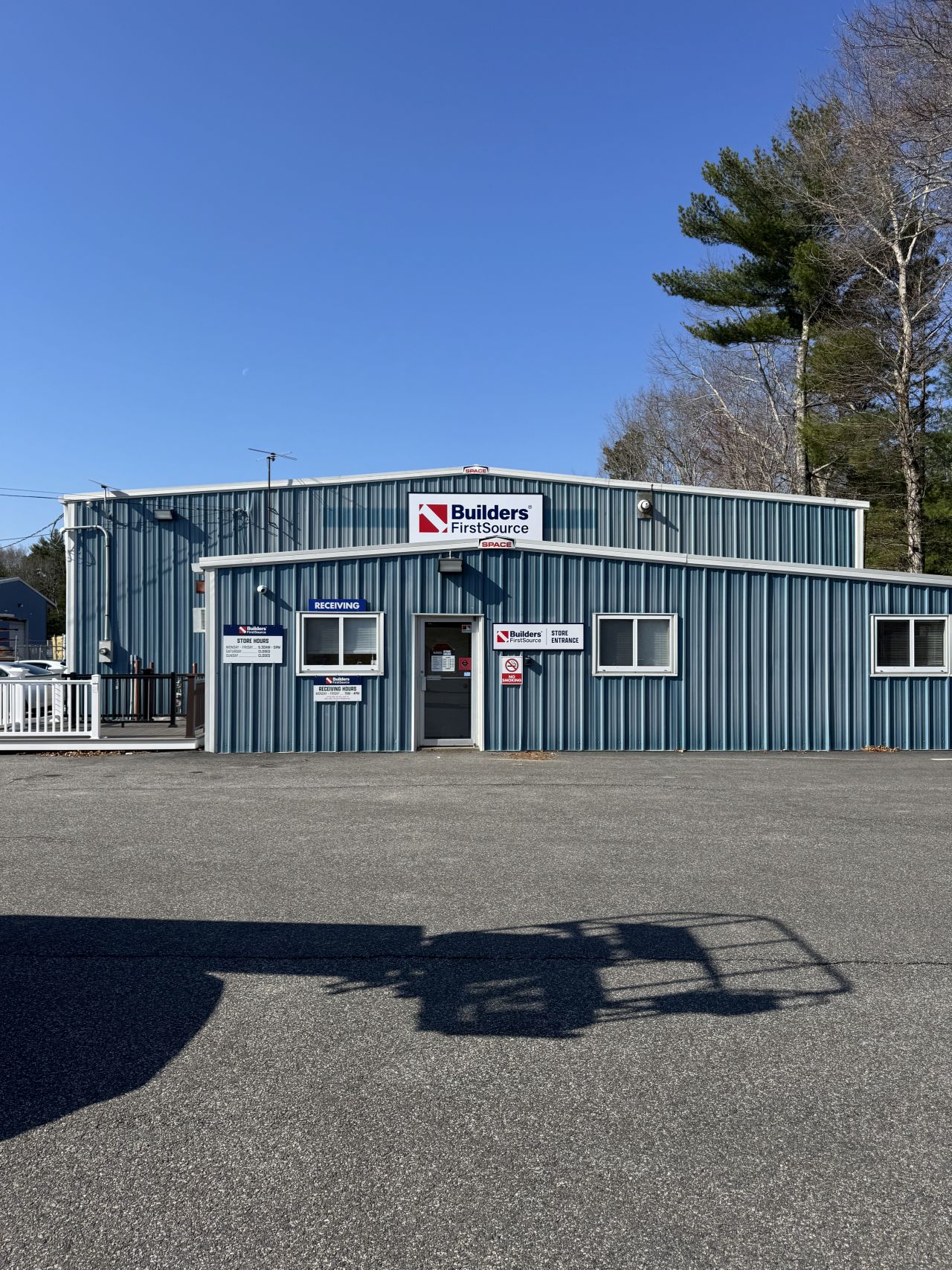 Exterior view of a Builders FirstSource building materials store with blue metal siding, front entrance doors, company signage, and paved customer parking area under a clear sky.