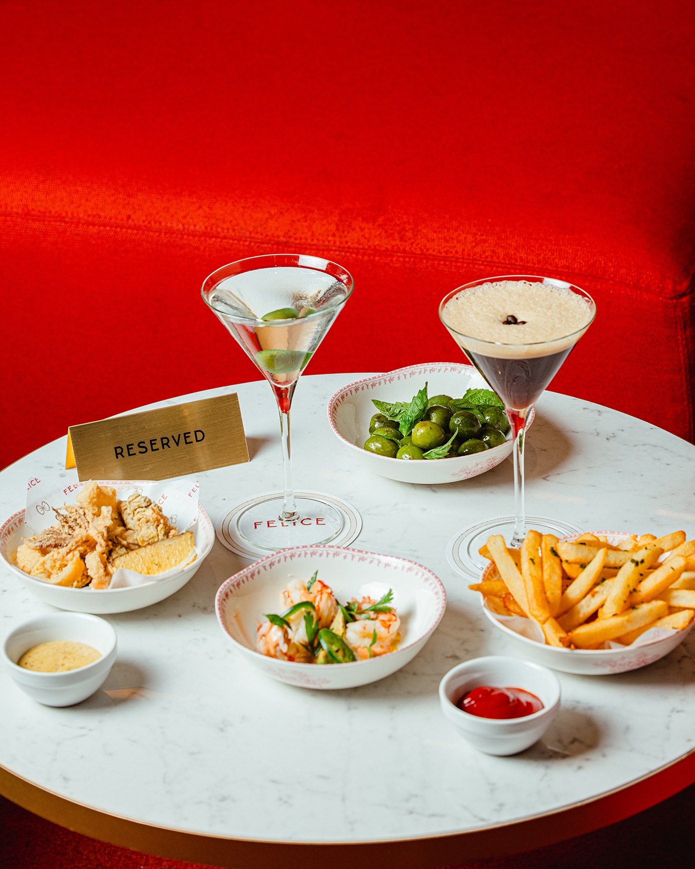 A marble cocktail table with a "Reserved" sign, featuring espresso martinis, fried calamari, shrimp cocktail, and fries against a vibrant red backdrop.