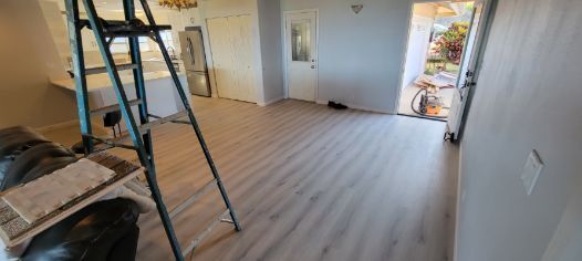 A recently renovated room with light gray laminate flooring and freshly painted light gray walls. A metal ladder stands in the foreground, with a kitchen and a glass-paneled door visible.