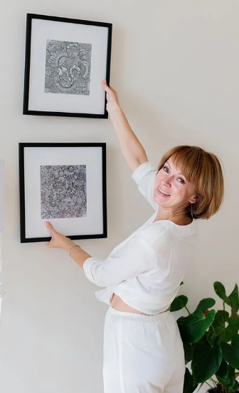 A happy senior woman with a short bob haircut, wearing casual white clothing, is shown in the process of hanging a second black-framed square picture on a plain wall, above an existing one. She is looking back over her shoulder at the camera with a cheerful expression.