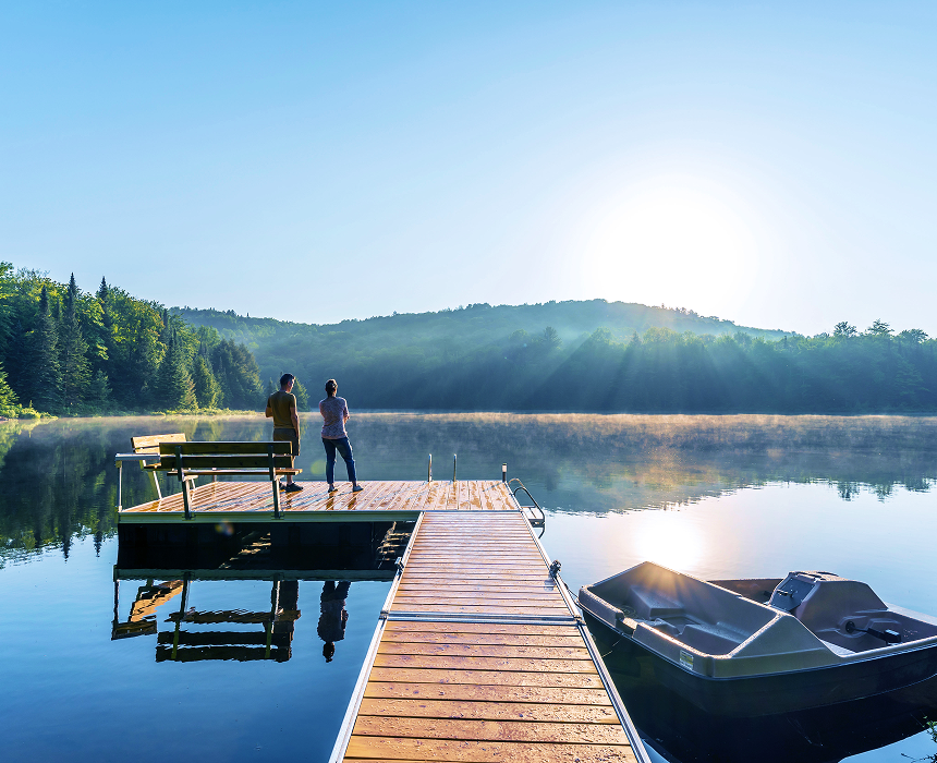Two individuals standing on a dock overlooking a calm lake at golden hour.