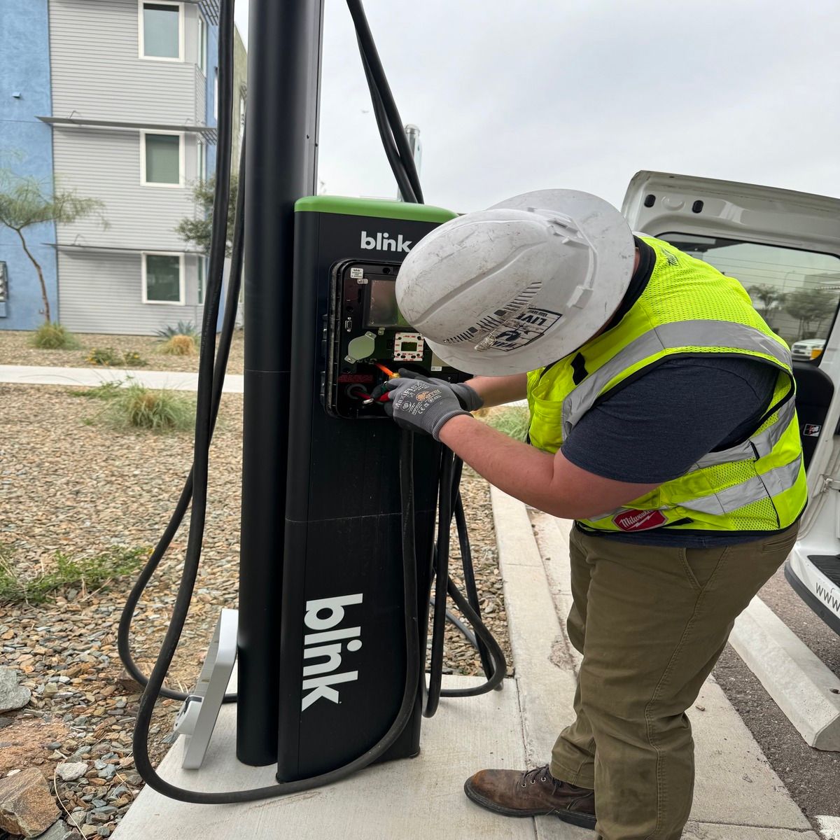 An electrician wearing a hard hat and high-visibility vest services an open Blink electric vehicle charging station beside a parking area, working carefully on internal wiring and electrical components outdoors.