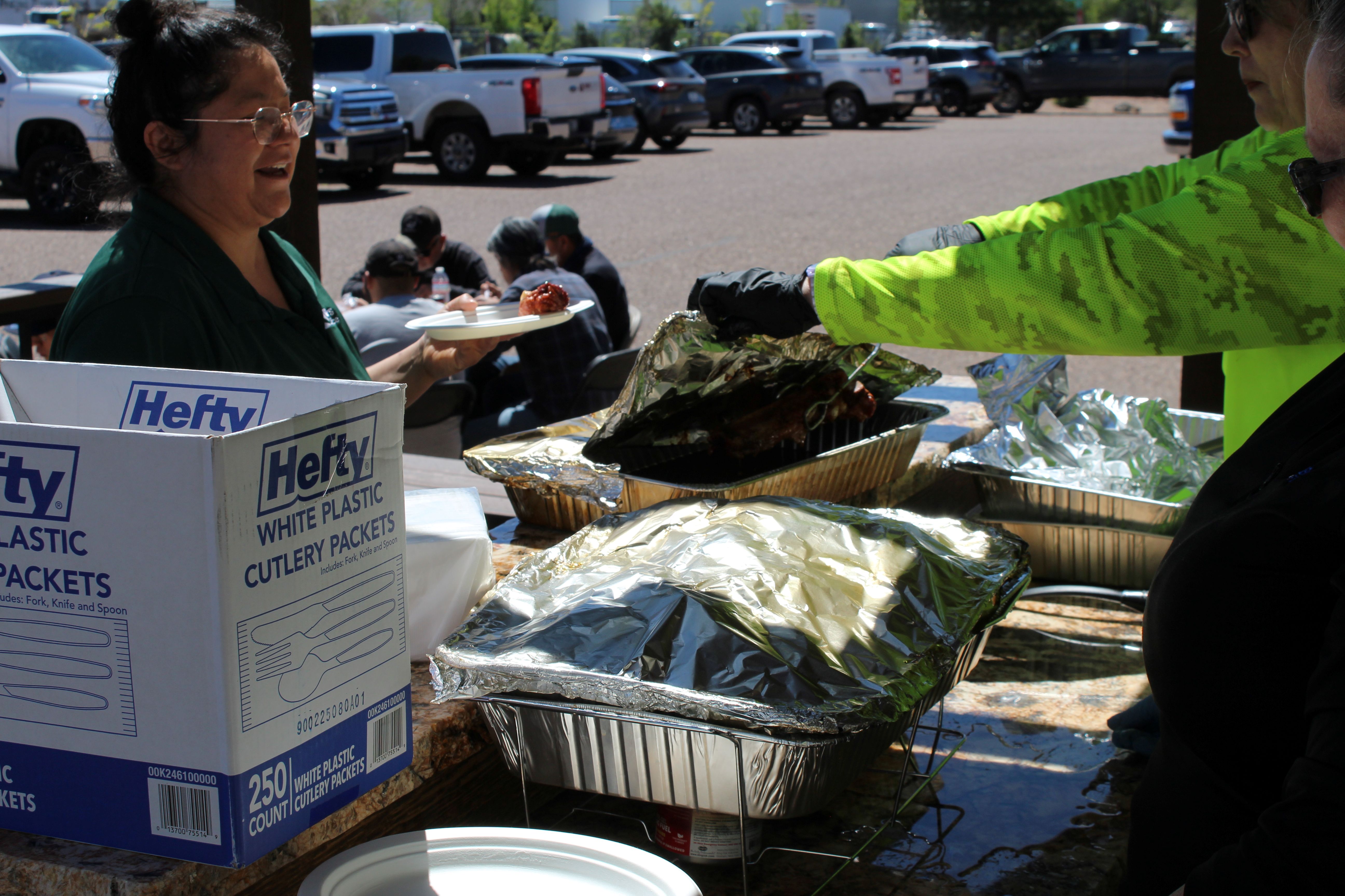 Outdoor food-serving station with foil-covered trays and a grill; one person uses tongs to serve food while another person holds a plate, with boxes of disposable cutlery on the counter.
