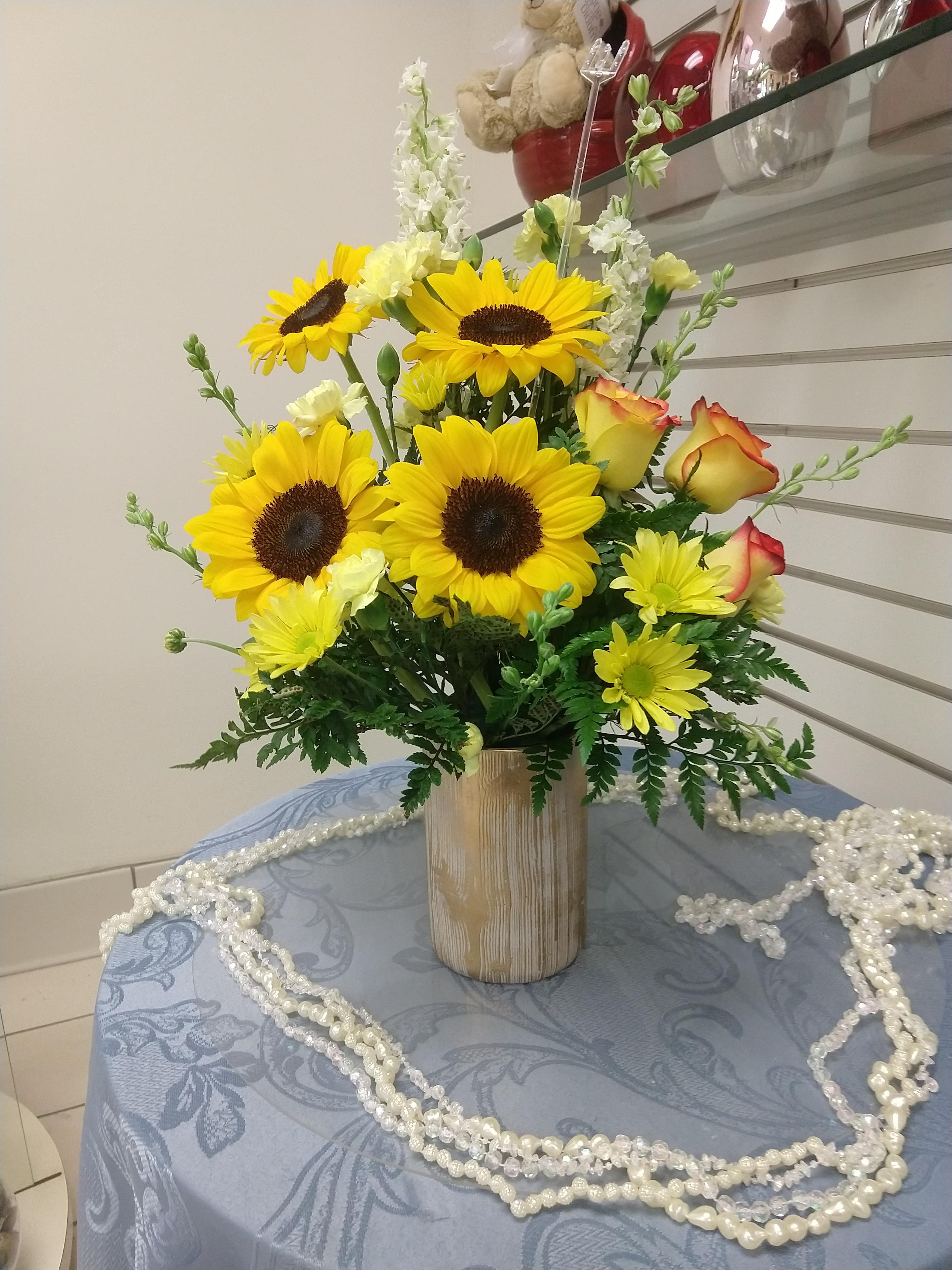 A burlap cylinder container filled with pretty sunflowers, orange spray roses, yellow daisies, and foliage.