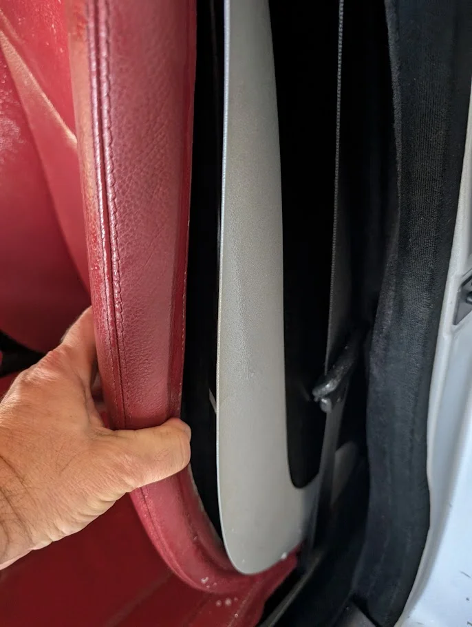 A hand is seen gripping a red leather car seat. The view is from inside a vehicle, looking towards the door frame. The seat appears to be a driver's or passenger's seat. The interior of the car features black fabric and trim, and a light gray plastic trim piece is visible near the door opening. The overall impression is of the interior of a car, possibly being inspected or adjusted.