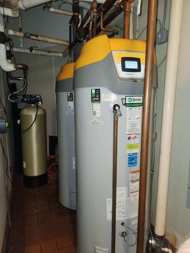 Two large, grey A.O. Smith Cyclone commercial water heaters with yellow tops are installed in a utility room. The professional setup includes complex overhead copper piping and a water filtration tank.