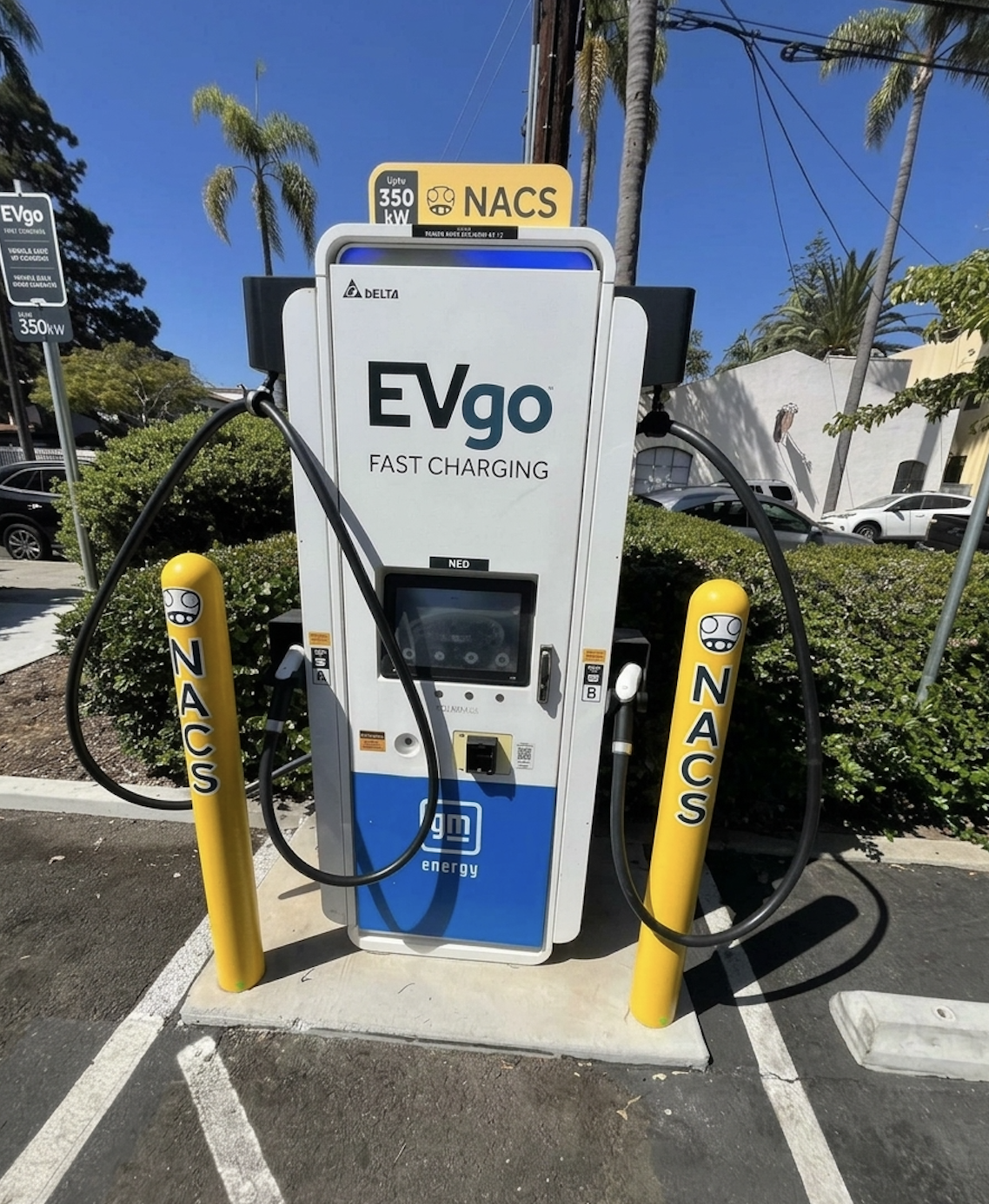 NACS EVgo fast charging unit installed in marked parking space at a public charging station, with EVgo logos on the charger, and yellow protective bollards around the unit.