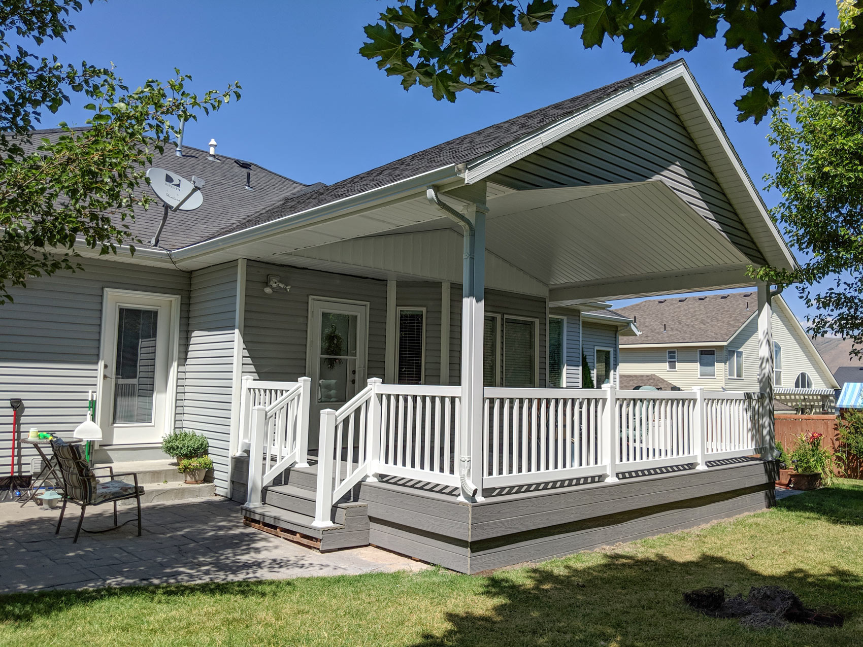 Create the perfect outdoor retreat with a custom covered composite deck and paver patio from DeckPro LLC. This design features low-maintenance grey composite decking and skirt boards, seamlessly integrated under a high-pitched gabled roof cover. The clean white railings and stairs provide a safe, defined space, making it easy to transition from the cozy, shaded deck to the open paver patio and lush backyard.