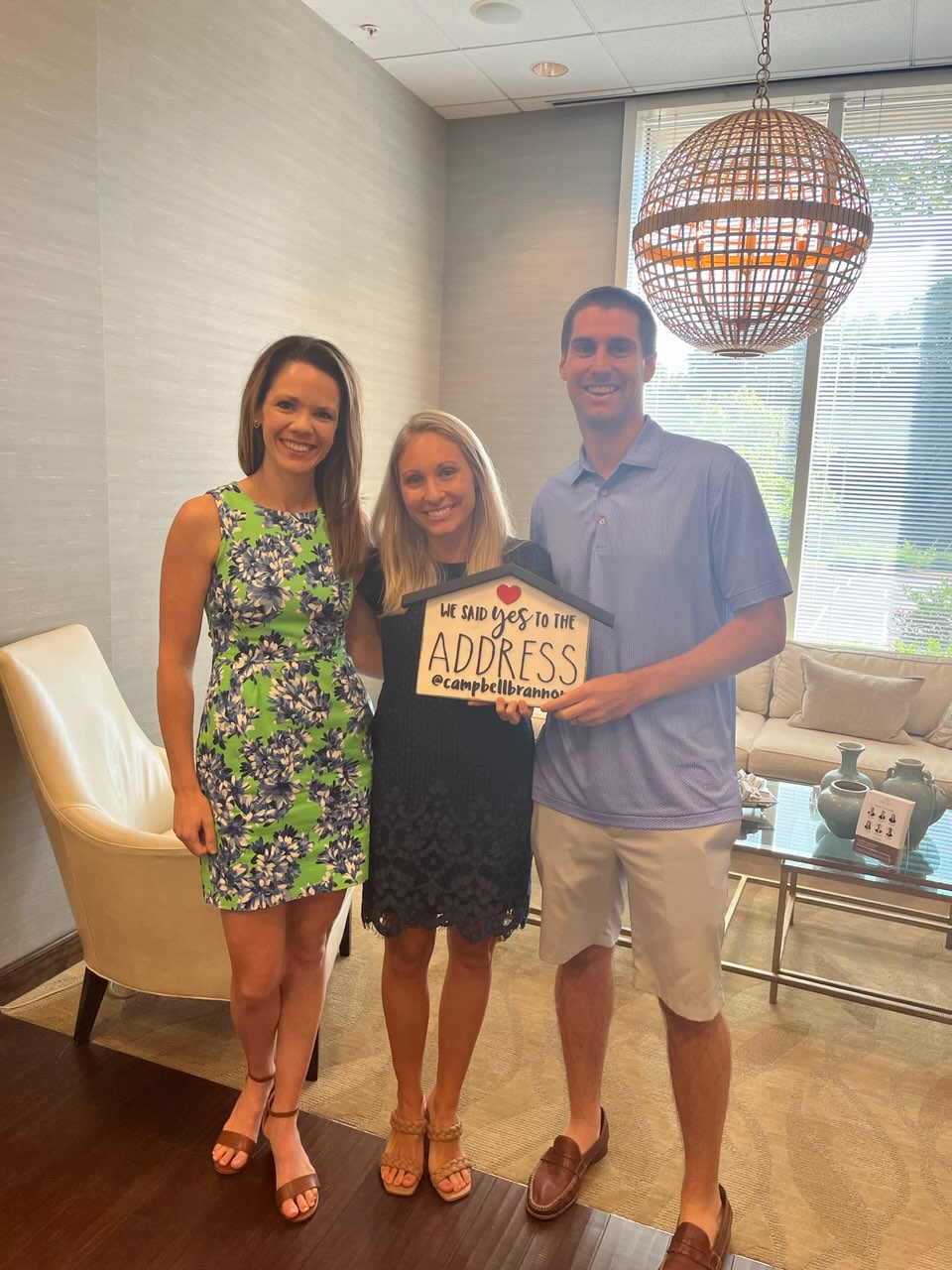 Three smiling people are standing together in a well-lit room, holding a decorative sign. The woman on the left wears a vibrant green and blue floral dress and brown sandals. The woman in the middle wears a black lace dress and tan braided sandals, holding the sign. The man on the right is in a light blue polo shirt and beige shorts, with brown loafers. The sign they hold reads "WE SAID yes TO THE ADDRESS" with a small heart above the 'yes' and "@campbellbrannon" below. The background features a cream-colored armchair, a decorative spherical light fixture, and a sofa with decorative items on a glass coffee table.