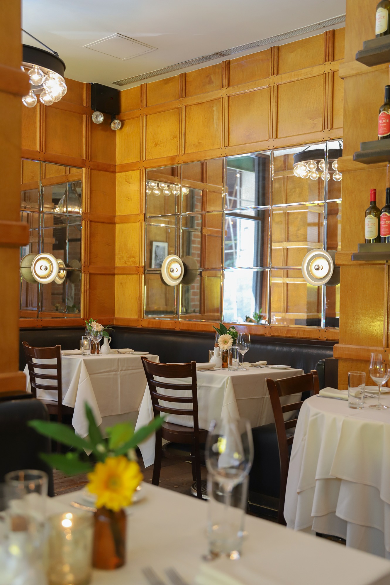 An elegant dining area features light wood walls with a grid of inset mirrors. Tables are set with white linens, glasses, and yellow flowers, creating a bright and sophisticated atmosphere.