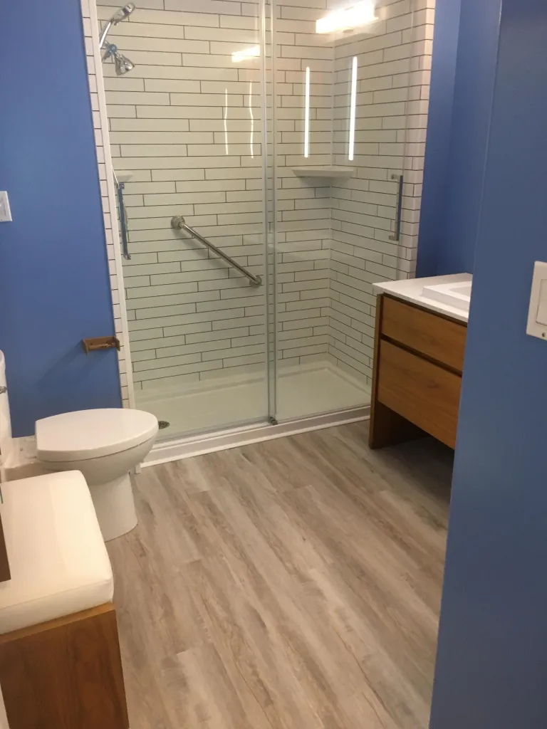 A modern bathroom with blue walls, a white toilet, a glass-enclosed shower with white subway tiles, and a wooden vanity with a white countertop.