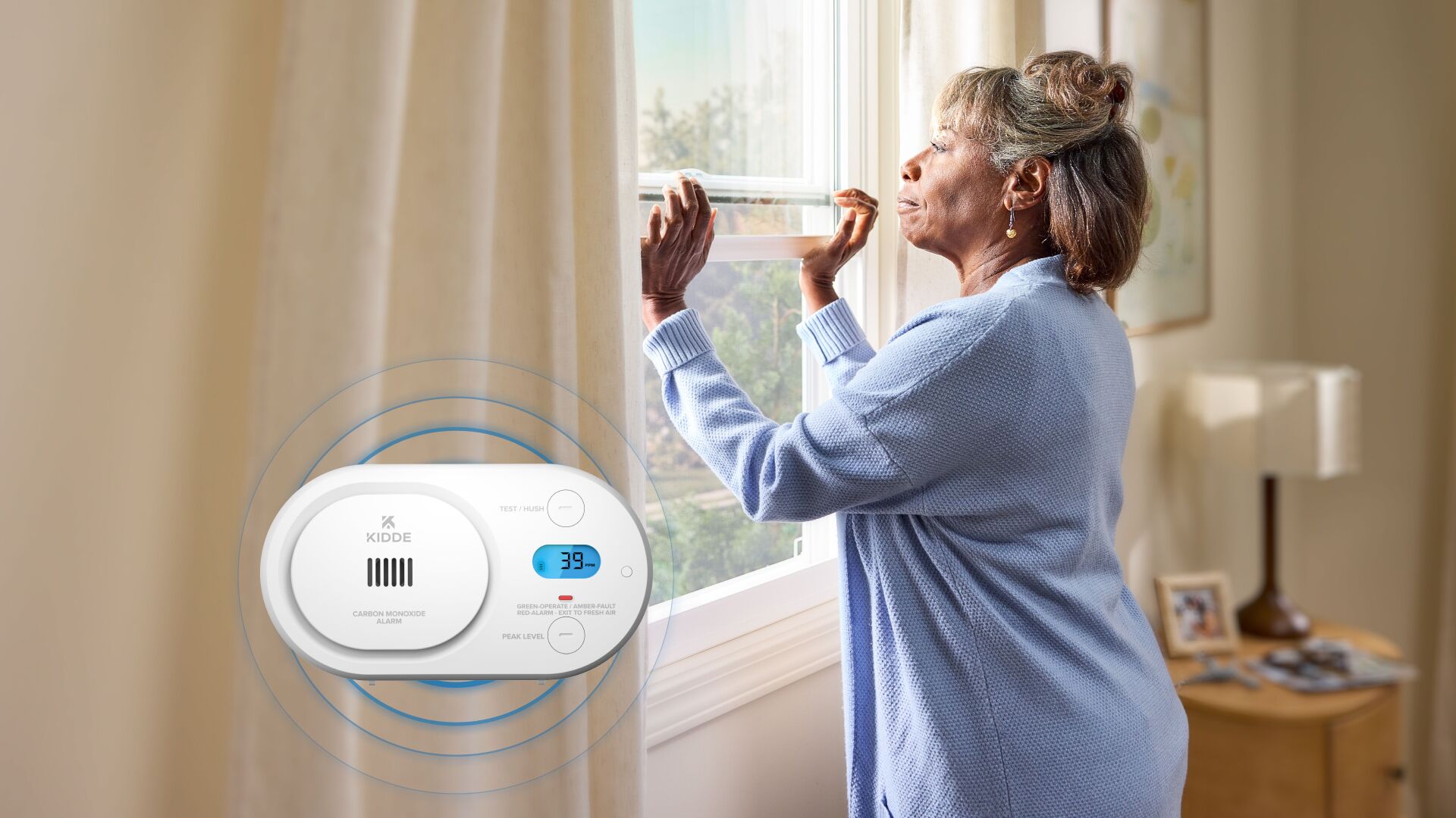 A woman opens her window. In the foreground, a carbon monoxide alarm is going off, showing that 39 ppm has been detected in the air. Blue circles representing the sound is emanating from the device.