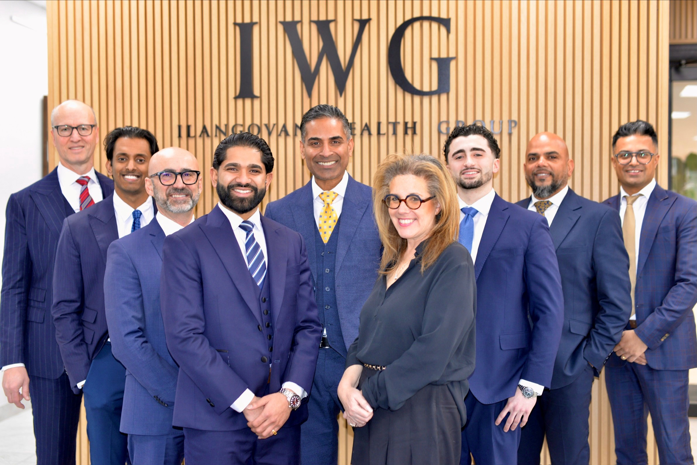 Professional team photo of the Ilangovan Wealth Group of RBC Dominion Securities. Nine team members in business attire stand confidently in front of a modern wooden slat wall featuring the IWG logo and company name. The group includes diverse professionals wearing navy and charcoal suits with colorful ties, positioned in their office setting.