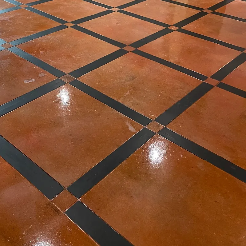 A close-up shot of a floor with a diamond pattern of reddish-brown tiles and black grout lines. The tiles have a glossy finish, reflecting light and creating bright highlights.