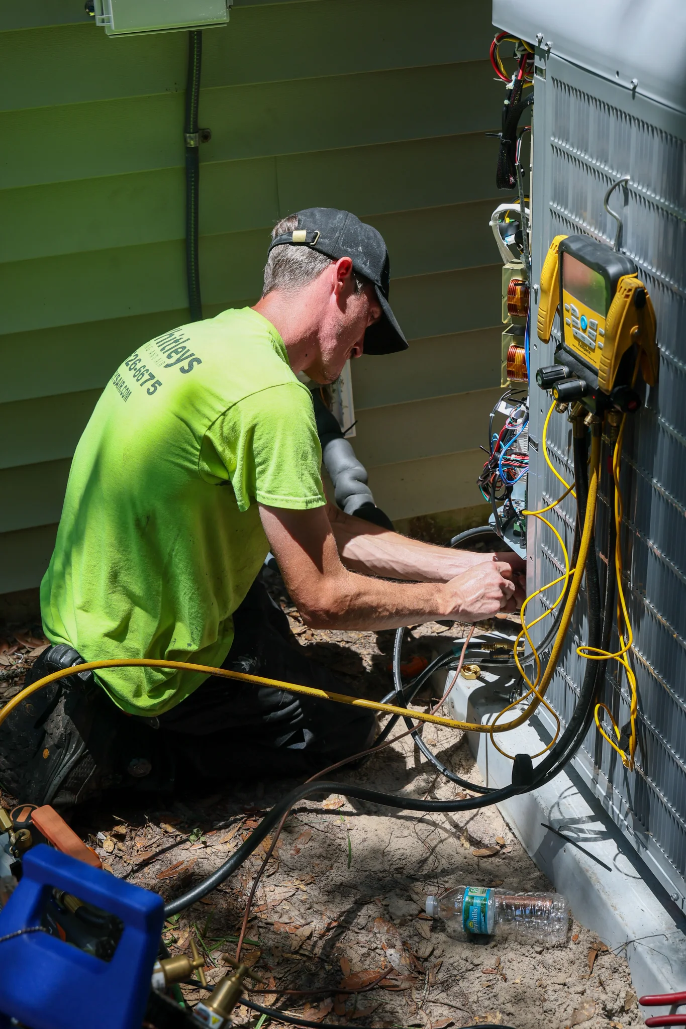 A man in a bright green shirt and black cap is working on an outdoor air conditioning unit. He is kn 