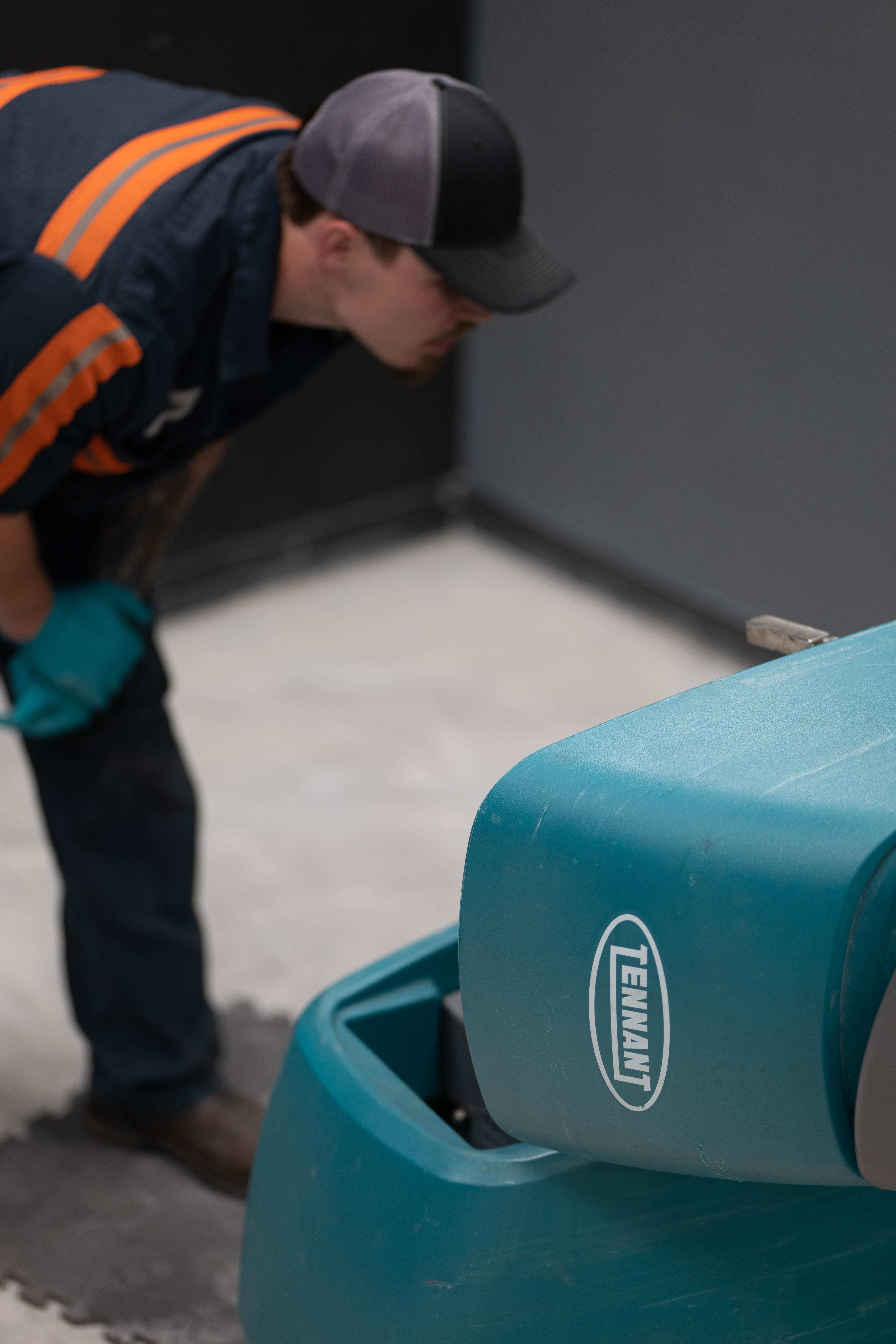 A WAY Solutions technician visually inspecting the lower chassis and squeegee mechanisms of a teal Tennant commercial floor scrubber inside a facility shop.