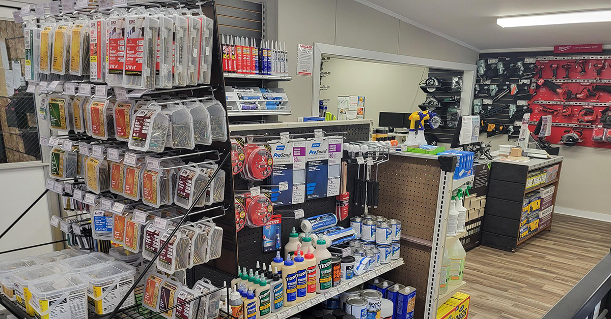 Interior showroom display of GRK wood screws and fasteners on a pegboard wall, with adhesive tubes, sandpaper rolls, and Titebond wood glue on adjoining shelves, opening into a checkout and tool display area in the background.