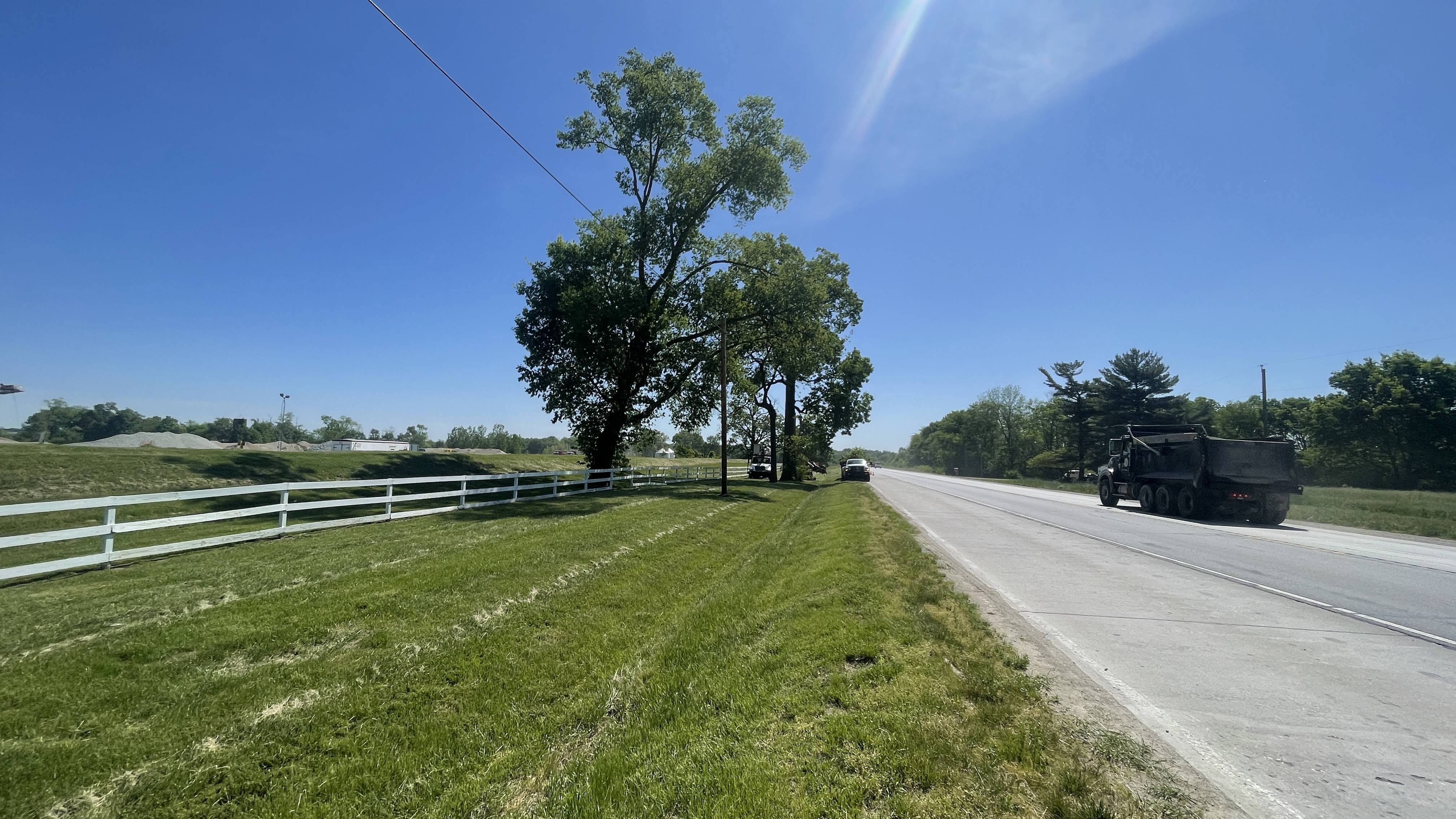 A dump truck drives down a paved road next to a grassy field with a white fence. Three large trees stand in the field. The sky is a clear blue with a rainbow-like lens flare from the sun.