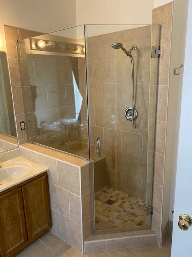 A modern neo-angle shower enclosure with glass panels and a chrome shower head and handle is shown in a bathroom. The shower features tiled walls and a patterned tiled floor. Adjacent to the shower is a vanity with a sink and wooden cabinets, and a mirror reflecting the shower and a window with blinds. The overall color scheme is neutral with beige and brown tones.