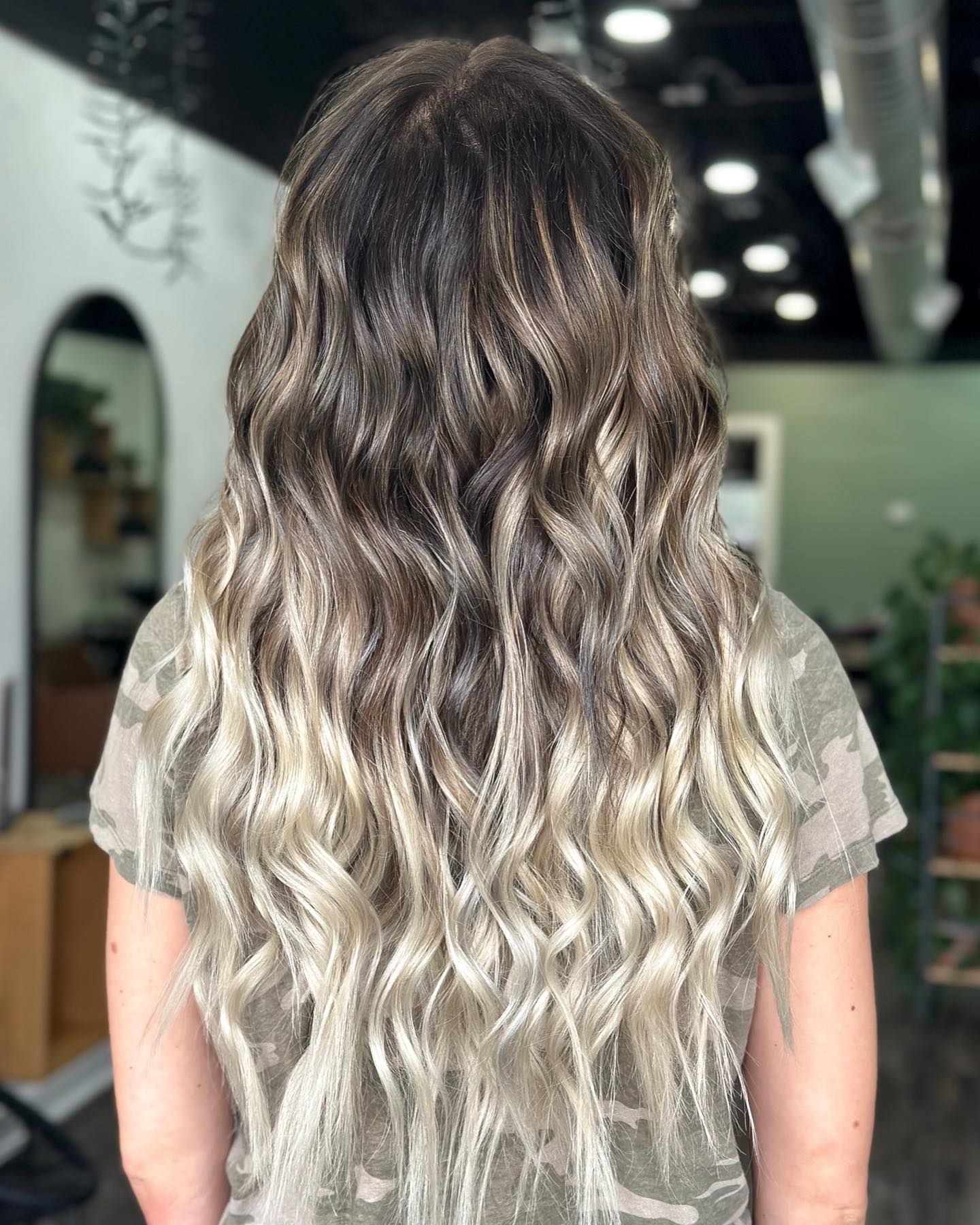 Back view of long wavy hair featuring dark brown roots blending into soft blonde balayage, styled professionally in a modern salon.