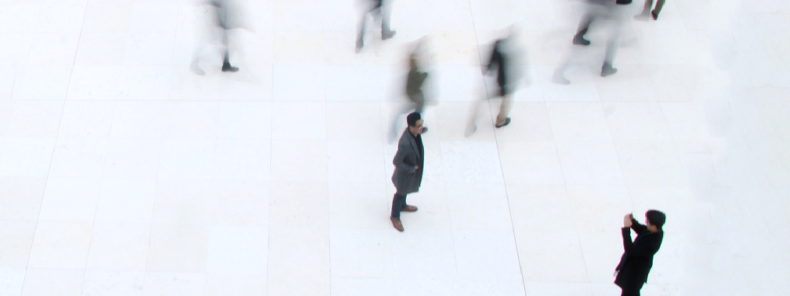 Overhead view of a man standing still in a busy public space as blurred figures walk past him.