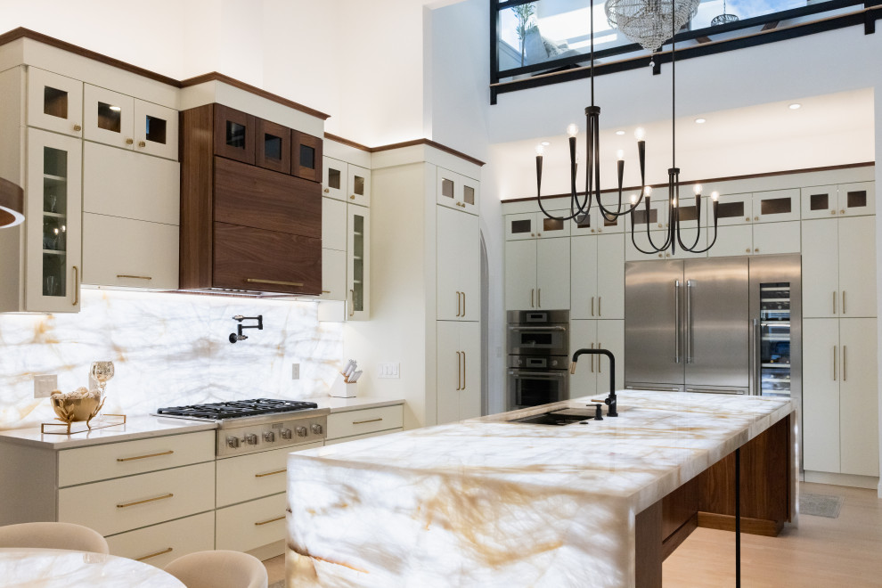 A luxurious, modern kitchen with a large island featuring a backlit onyx countertop. The kitchen has cream-colored cabinets, dark wood accents, stainless steel appliances including a double oven and a large refrigerator, and a contemporary black chandelier hanging from the high ceiling.